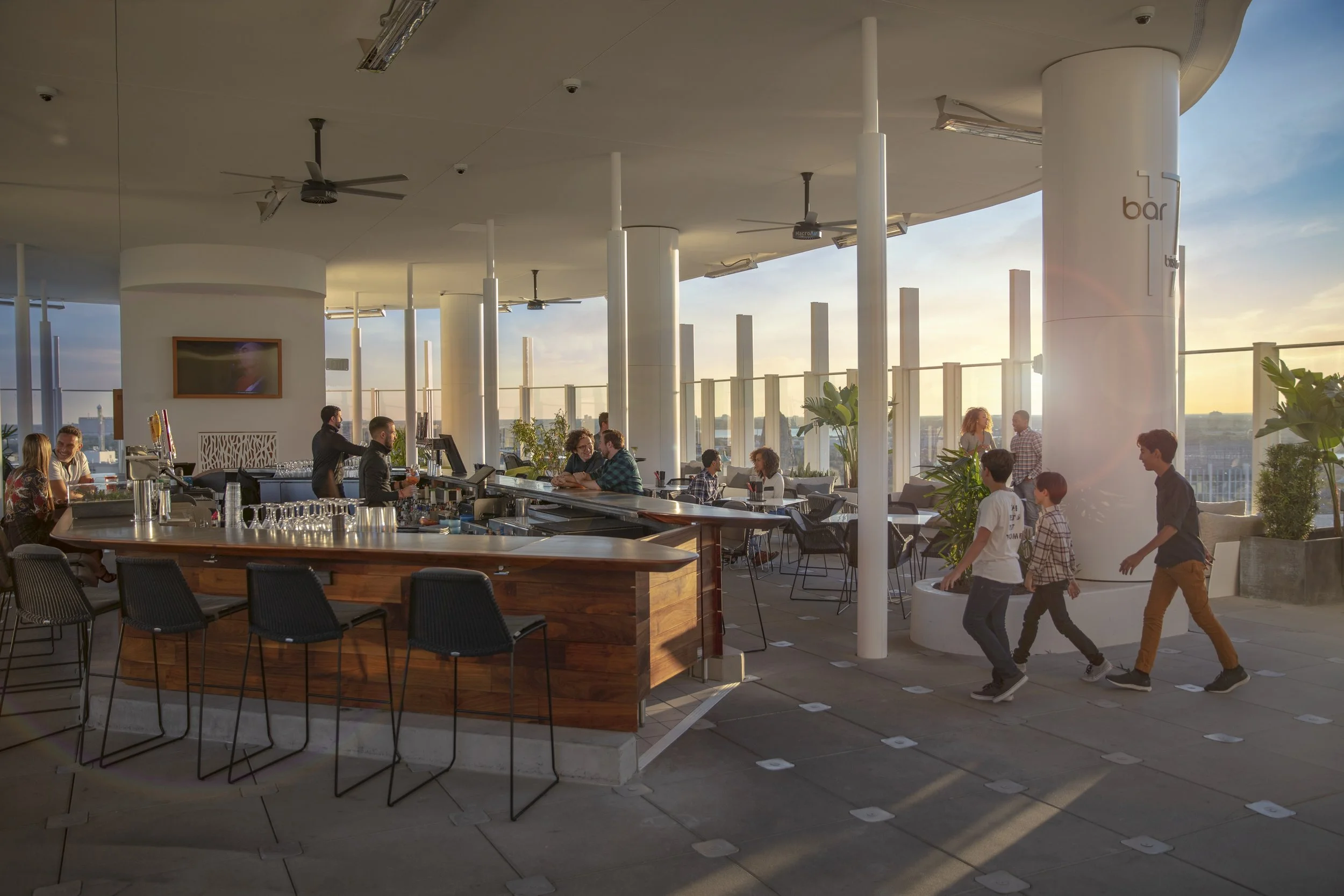 Indoor rooftop bar with people socializing, standing and sitting by the bar, and children walking in line. Large windows with a city view during sunset, green plants, ceiling fans, and a television on the wall.