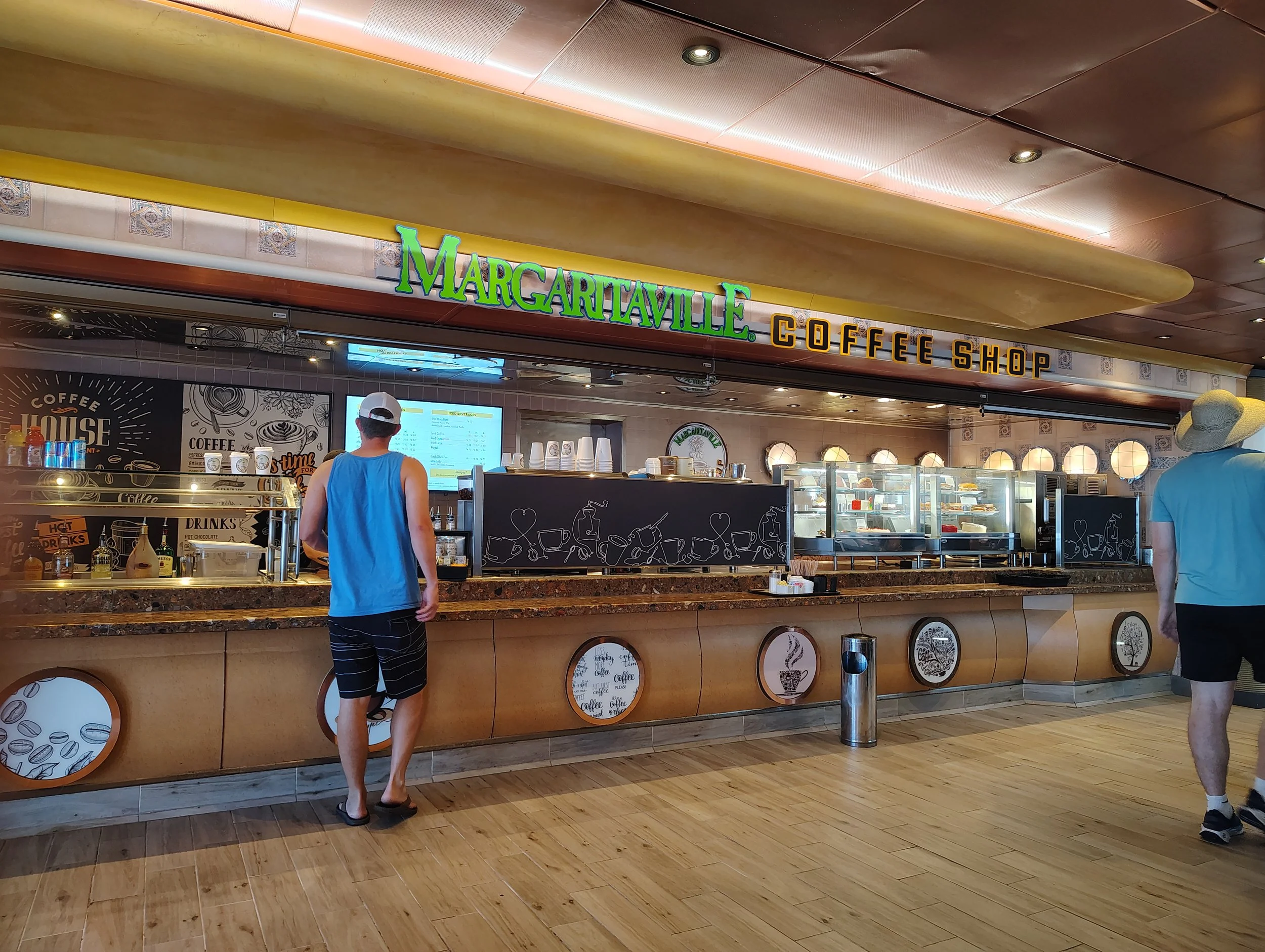 A coffee shop named 'Margaritaville Coffee Shop' with customers in line. The shop features a counter with display cases of food, a digital menu screen, and various coffee-related decorations.