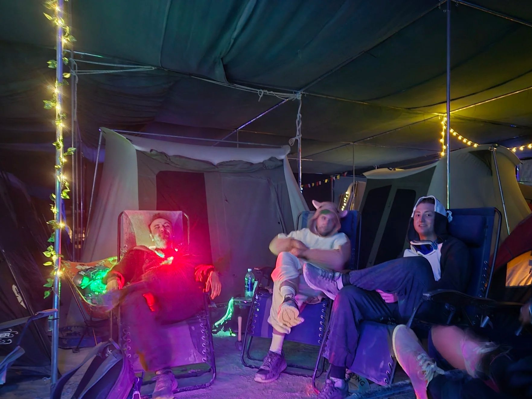 Four young adults relaxing in camping chairs inside a tent with colorful string lights at night, some wearing hoodies and hats, surrounded by tents.