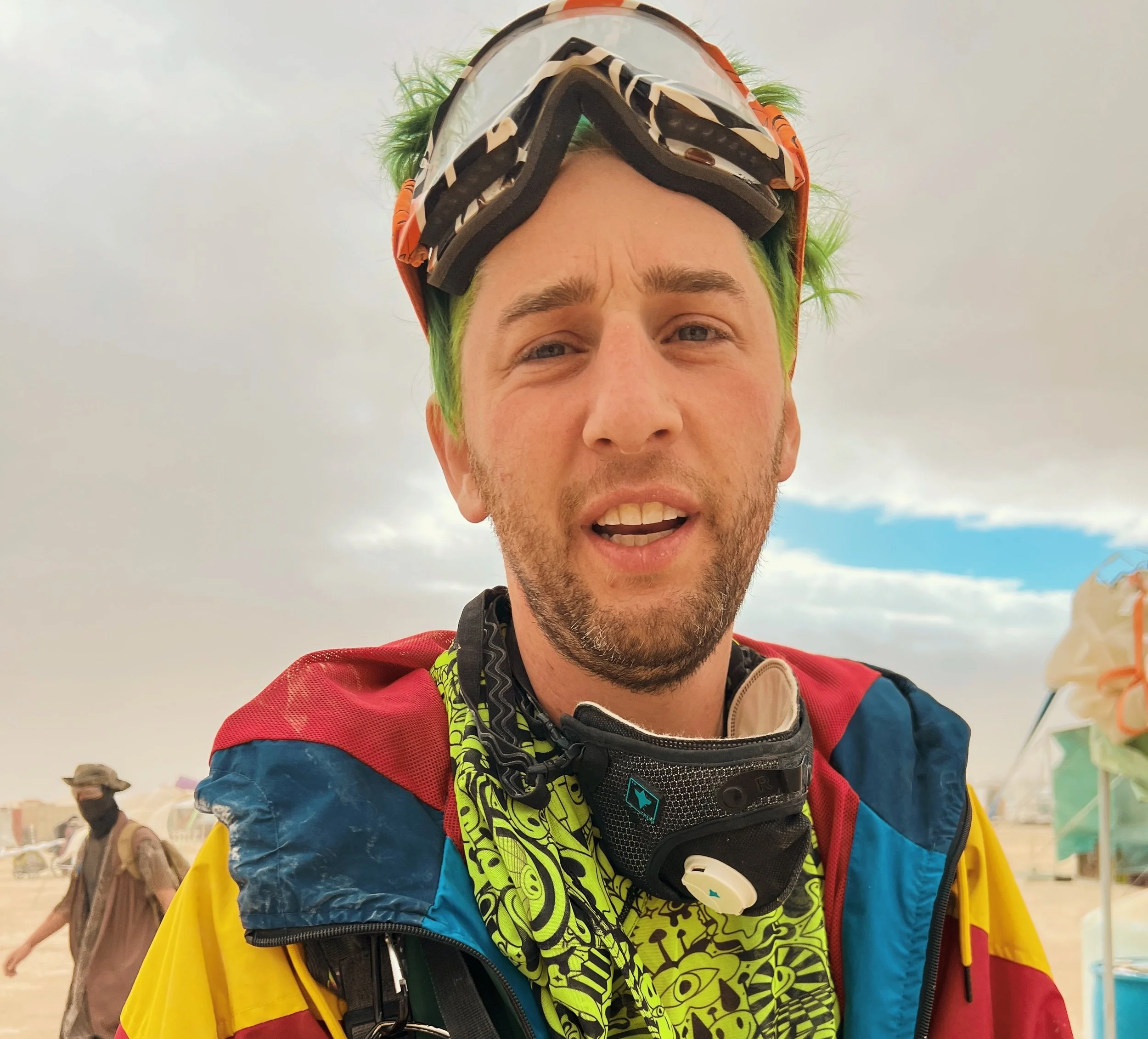 A smiling man with sunglasses, wearing a colorful cap, a patterned shirt open with no shirt underneath, yellow shorts, and accessories, standing outdoors at an event with bicycles and other people in the background.