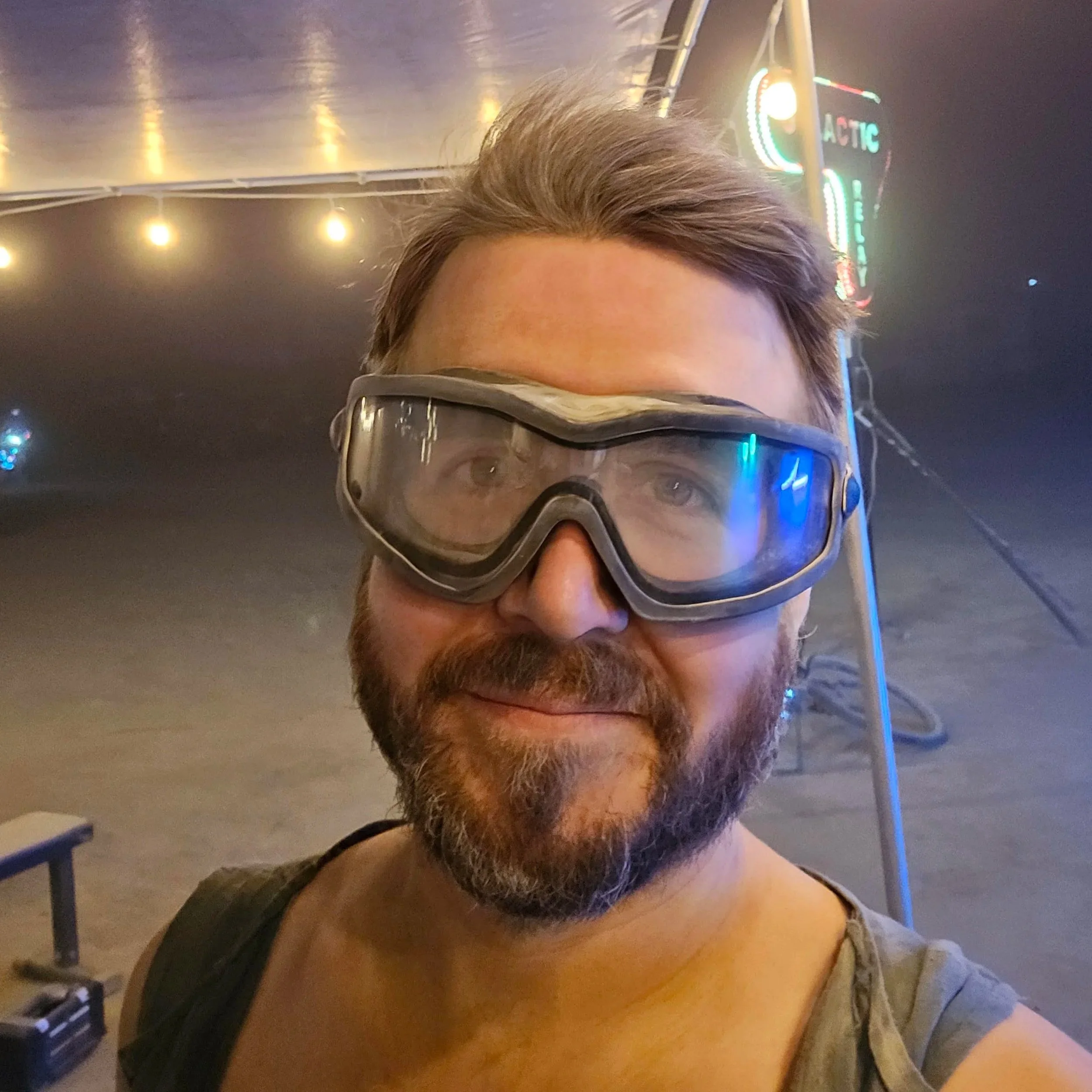 A man with a beard wearing protective goggles standing in front of a neon sign at night.
