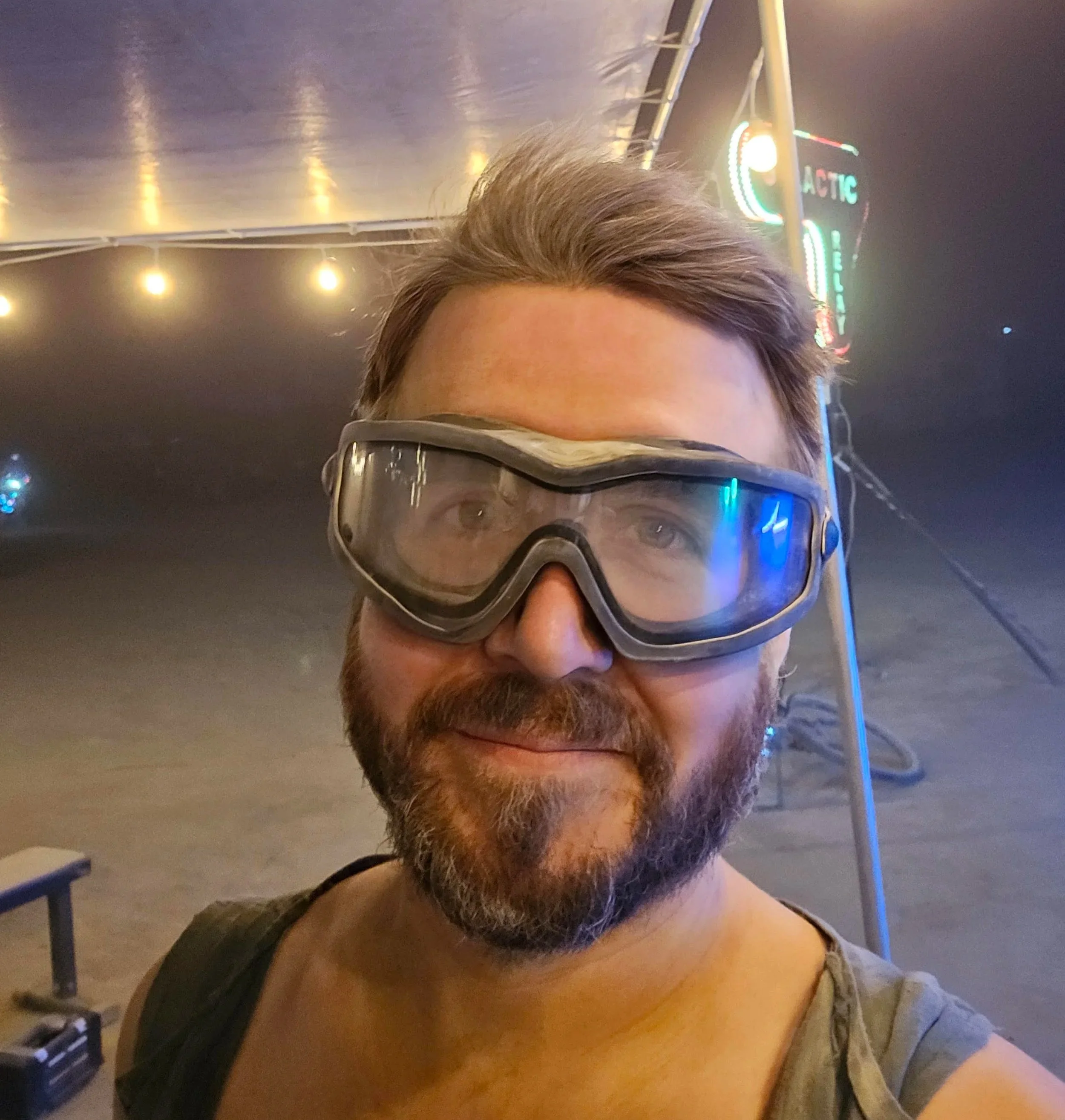 A man with a beard wearing protective goggles at an indoor ice skating rink, with neon lights and a sign in the background.