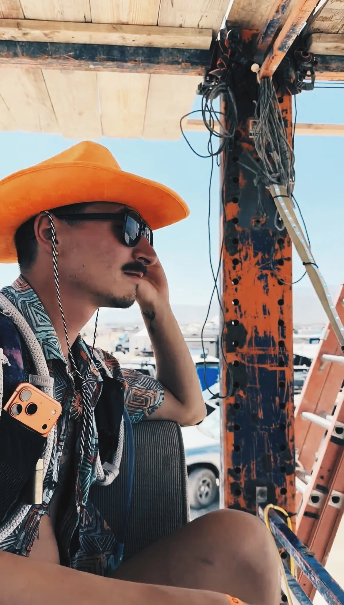 A man with a mustache and sunglasses sitting on a metal scaffold setup outdoors, wearing a colorful cap with floppy ears, a patterned shirt open to reveal a spiral tattoo on his chest, and holding onto the scaffolding with one hand. The background features a blue sky with some clouds.