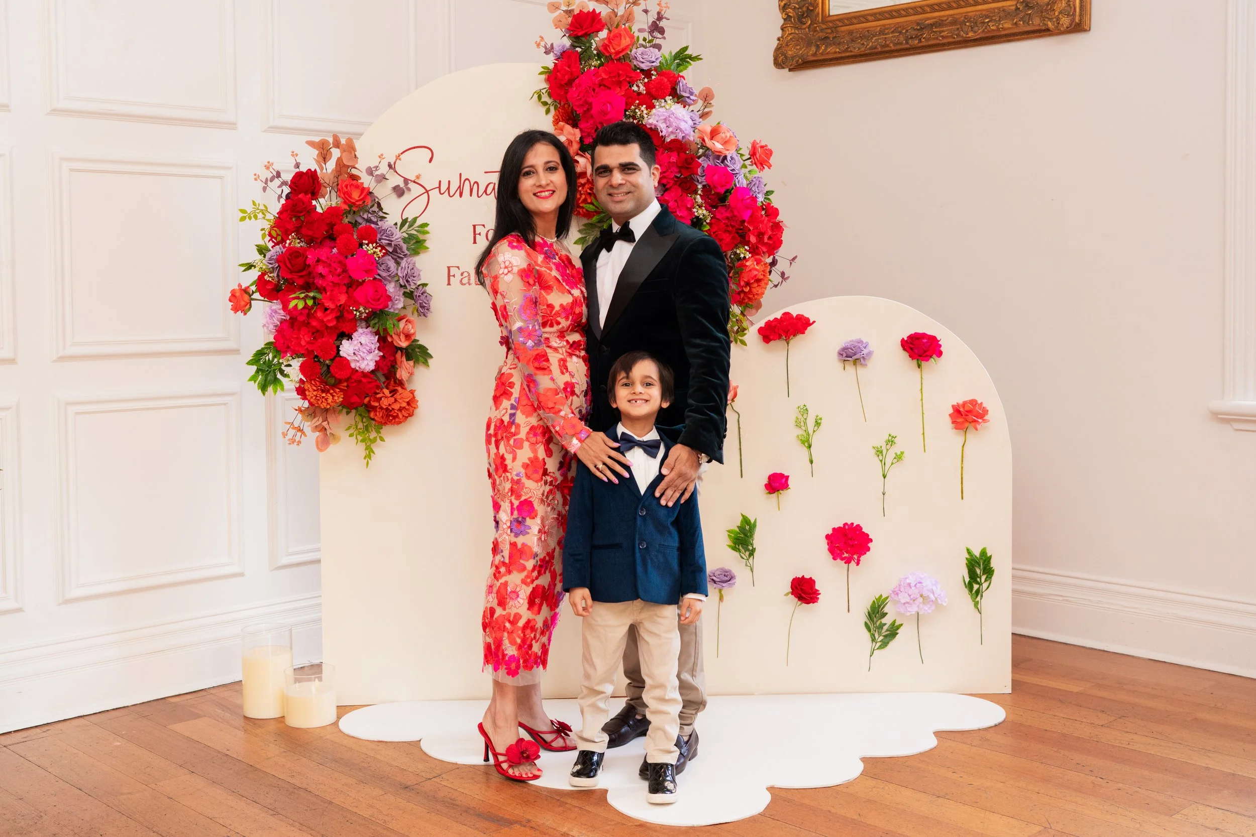A family of three stands in front of a flower wall at a celebration, smiling at the camera. The woman wears a floral dress, the man is in a tuxedo, and the child is dressed in a suit with a bow tie.