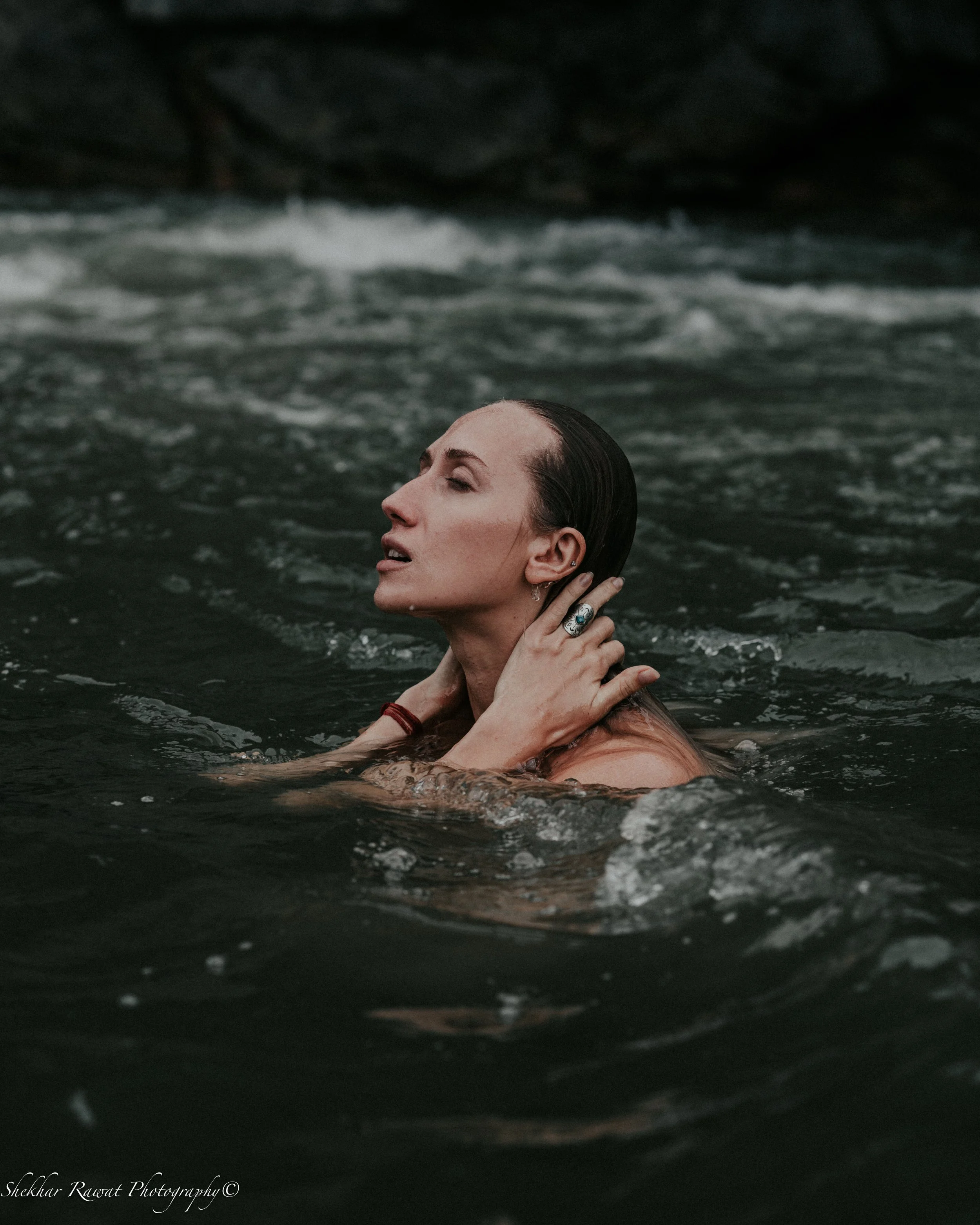 A woman with wet hair and closed eyes partly submerged in water, with her hands touching her neck, surrounded by dark water and a blurred background.
