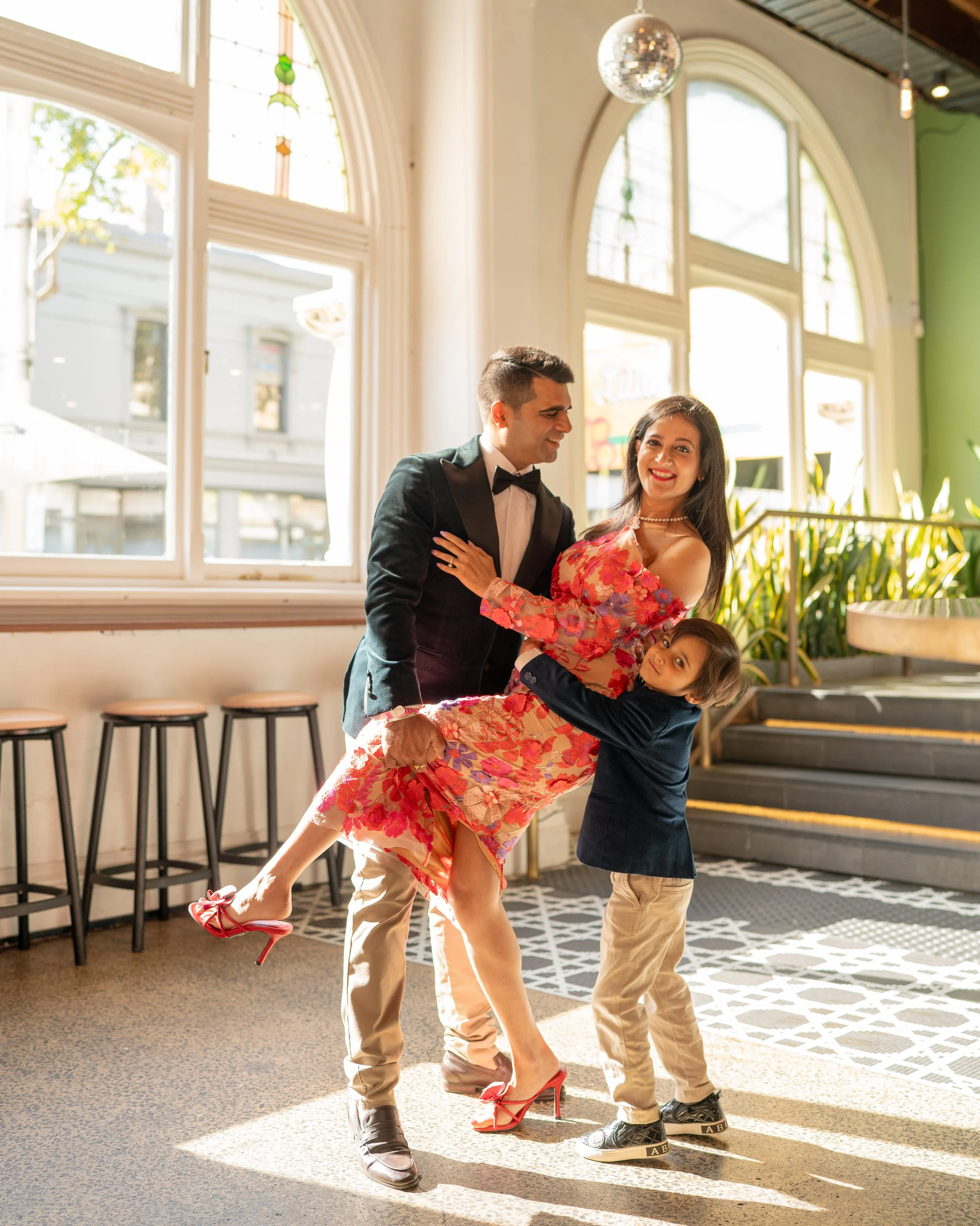 A man in a tuxedo and a woman in a floral dress with red high heels are dancing together with a young boy in a navy jacket and beige pants, indoors near large windows and a staircase, with natural light.