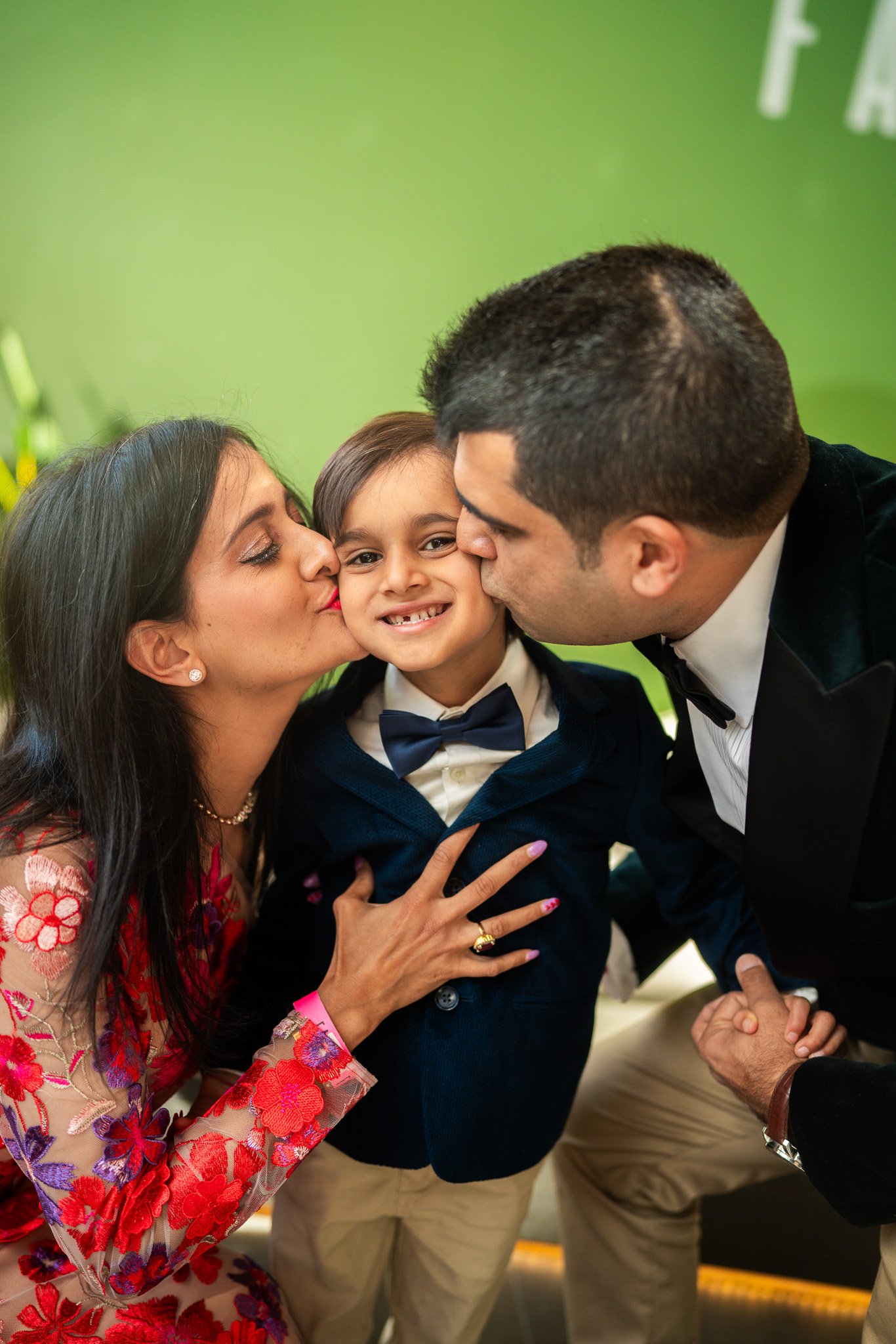 A woman and a man are kissing a young boy on the cheeks at a social event, with the woman wearing a colorful floral dress and the man wearing a dark suit and tie, against a green background.