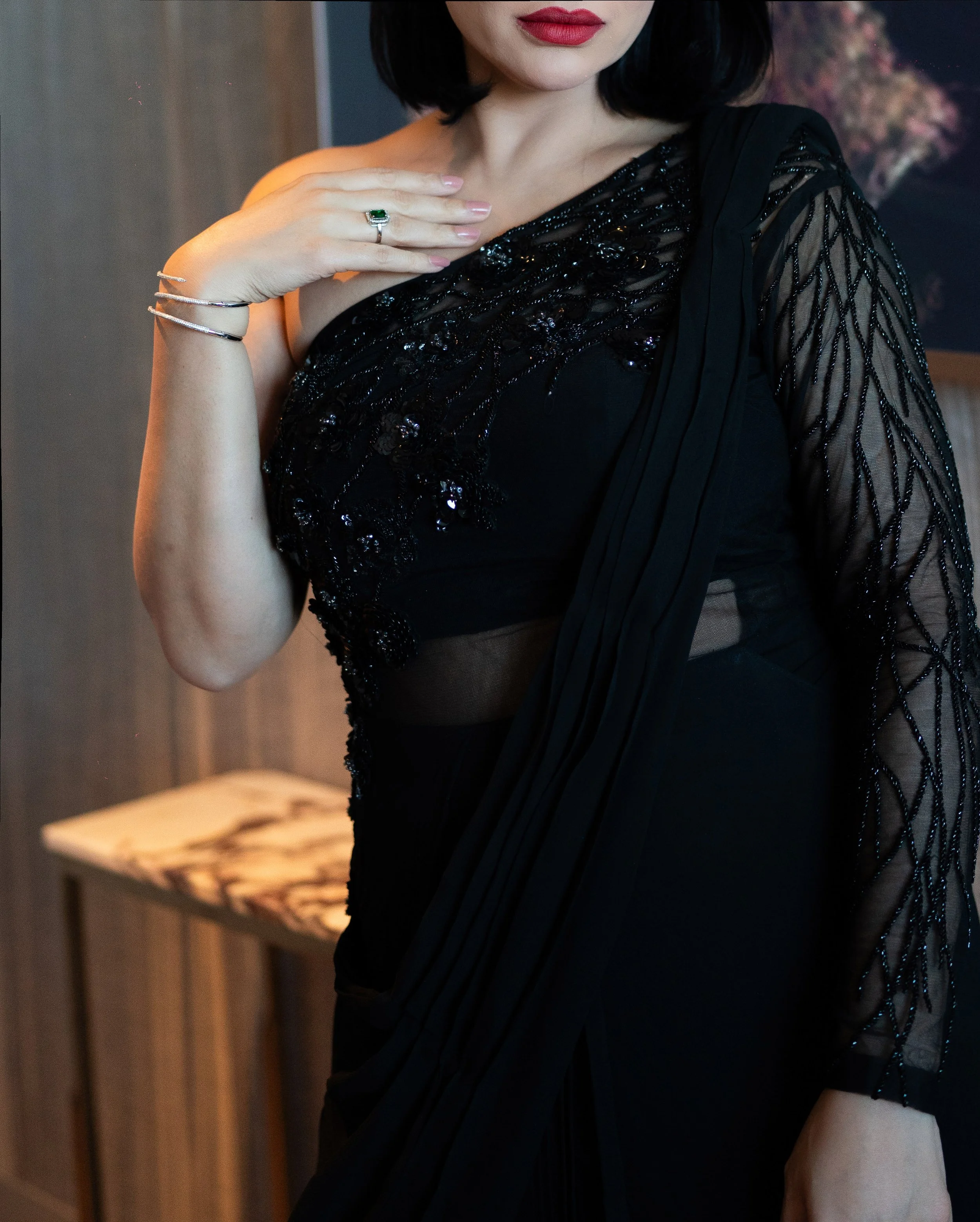 A woman with dark, shoulder-length hair wears a black dress with sheer and embroidered detailing, showing her shoulder and arm. She has red lipstick, a ring with a large green gemstone on her finger, and a silver bracelet on her wrist. She is touchin