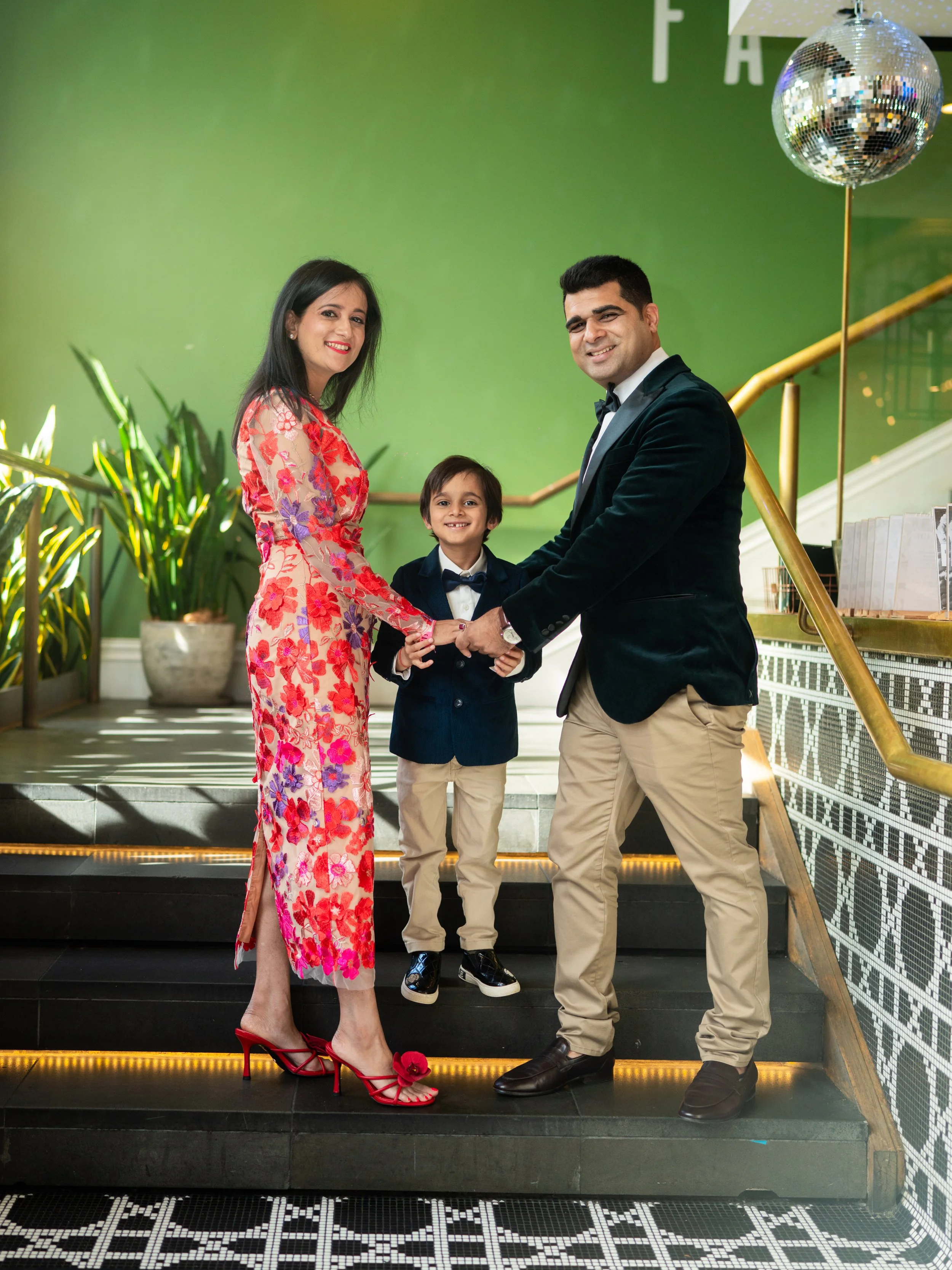 A family of three holding hands on a staircase inside a building with a green wall and a disco ball hanging from the ceiling. The woman is wearing a floral dress and red high heels, the man is in a tuxedo, and the boy is dressed in a navy blazer and 