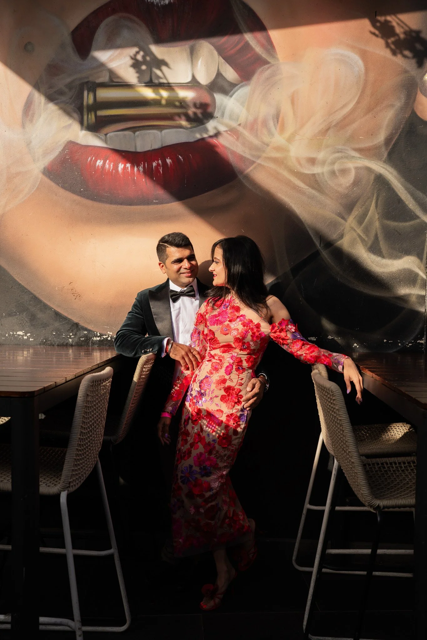 A man in a tuxedo and a woman in a pink and red floral dress pose together in front of a large mural of lips smoking a cigarette, with smoke swirling around the lips.
