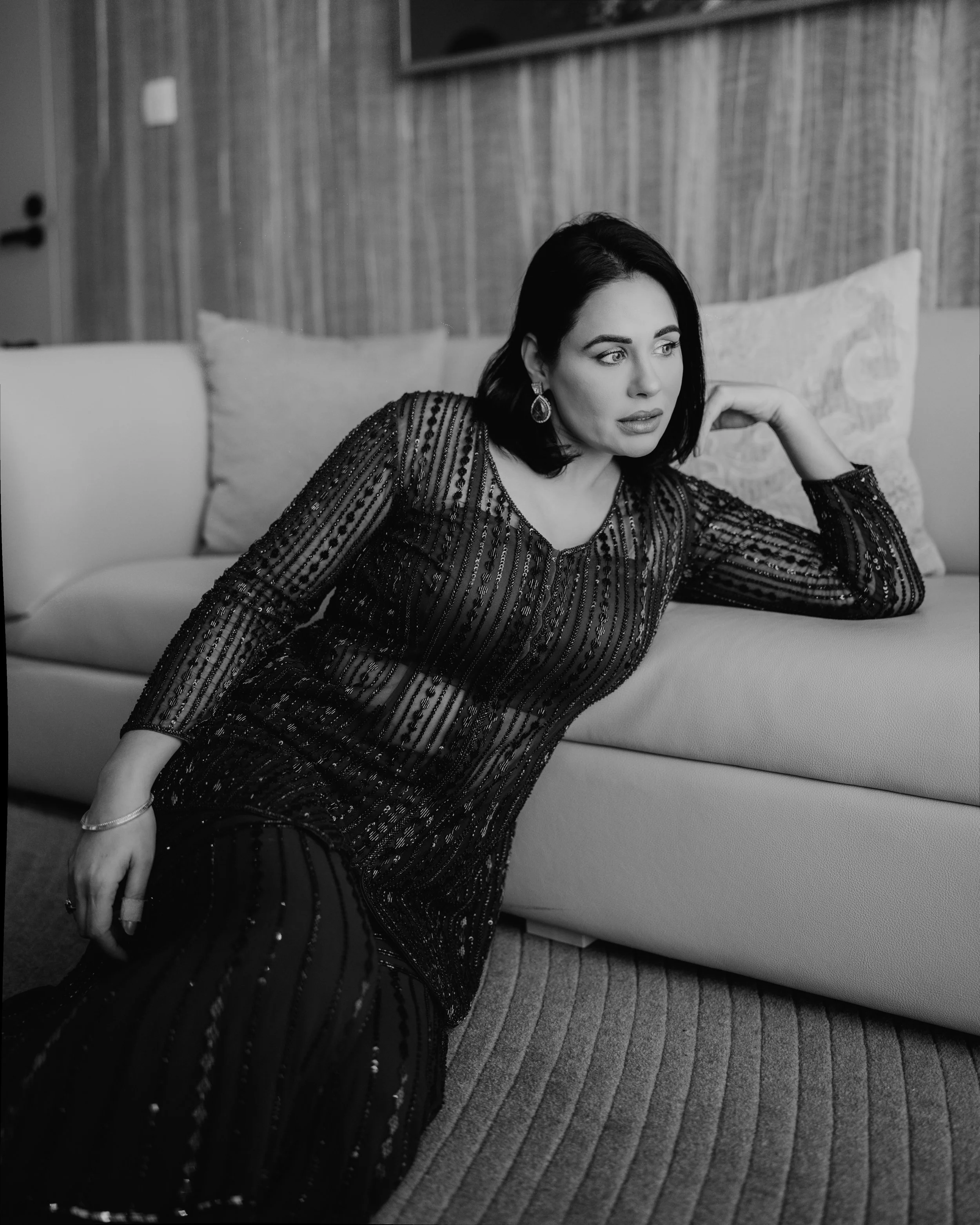 A black and white photo of a woman with dark hair, wearing a long-sleeved, beaded dress, sitting on a bench or sofa, with her head resting on her hand, looking thoughtfully to her right. The background features a wooden wall and some cushions or pill