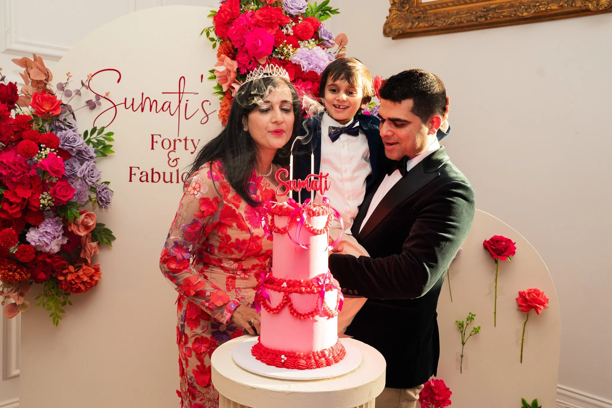 A woman wearing a tiara and floral dress, a man in a tuxedo, and a young boy in a suit are celebrating a birthday with a pink cake decorated with hearts and topped with candles. They are holding a cake knife together, and there is a backdrop of color