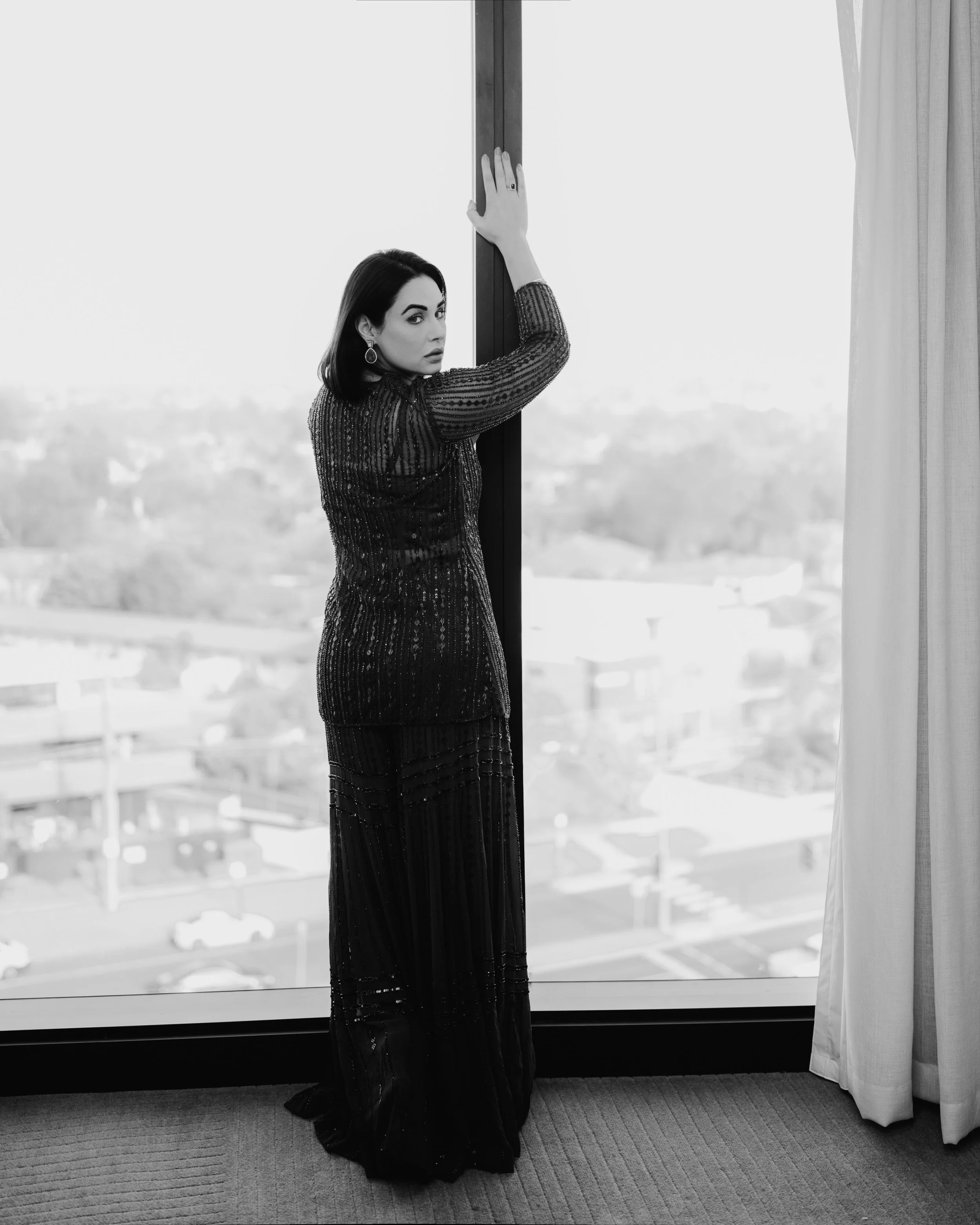 A woman in a long, elegant, beaded gown standing by a large window with a city view, looking over her shoulder at the camera, with her hand resting on the window frame.