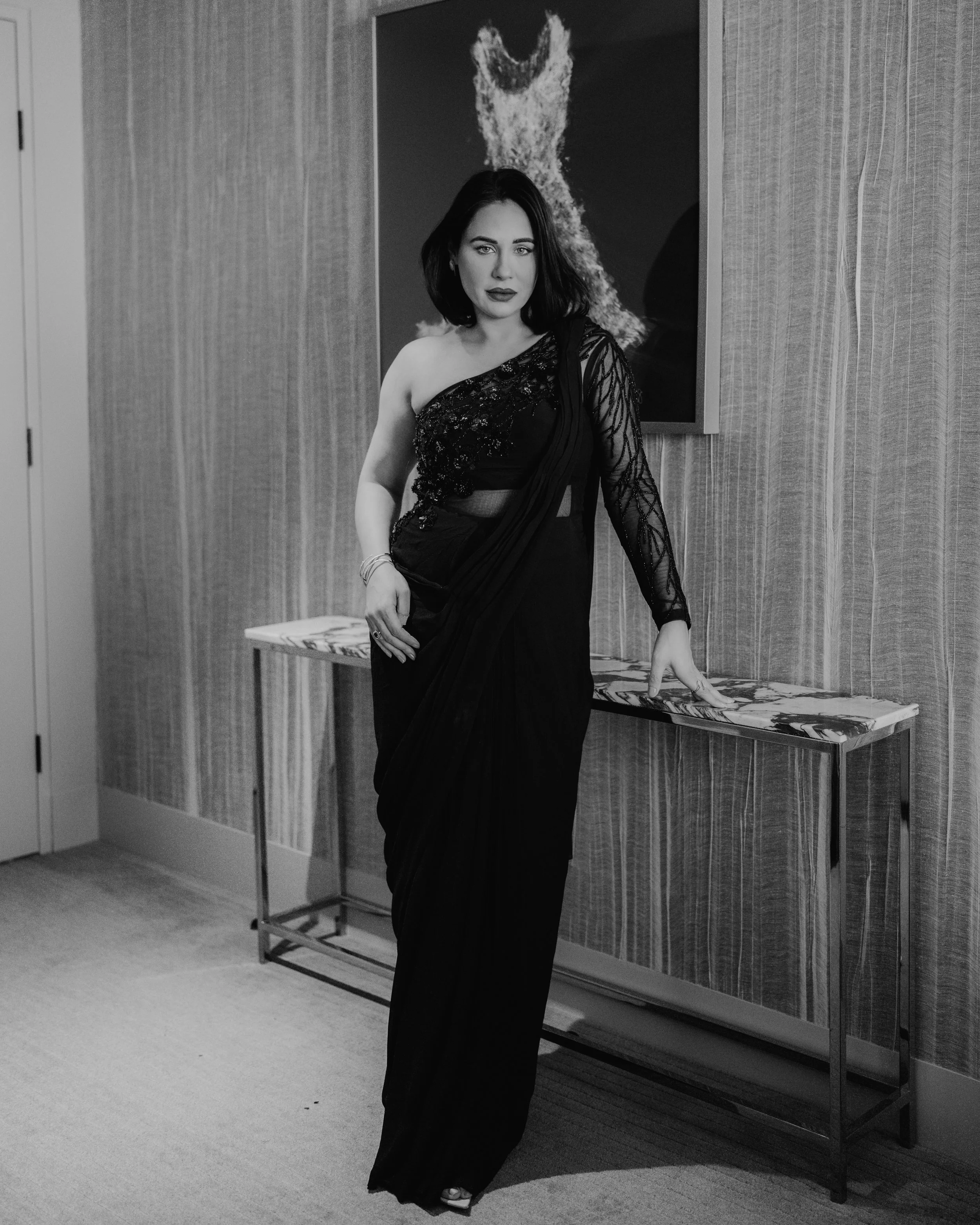A woman in a black dress standing indoors in front of a textured wall with a cow painting in the background.