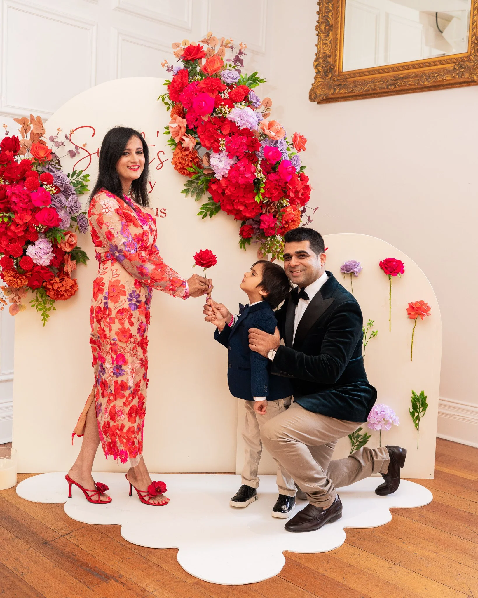A family of four, including a woman in a floral dress, a man in a tuxedo, and two children, are participating in a wedding ceremony. They are in front of a floral backdrop with red, purple, and pink flowers, and a white curved background with red wri