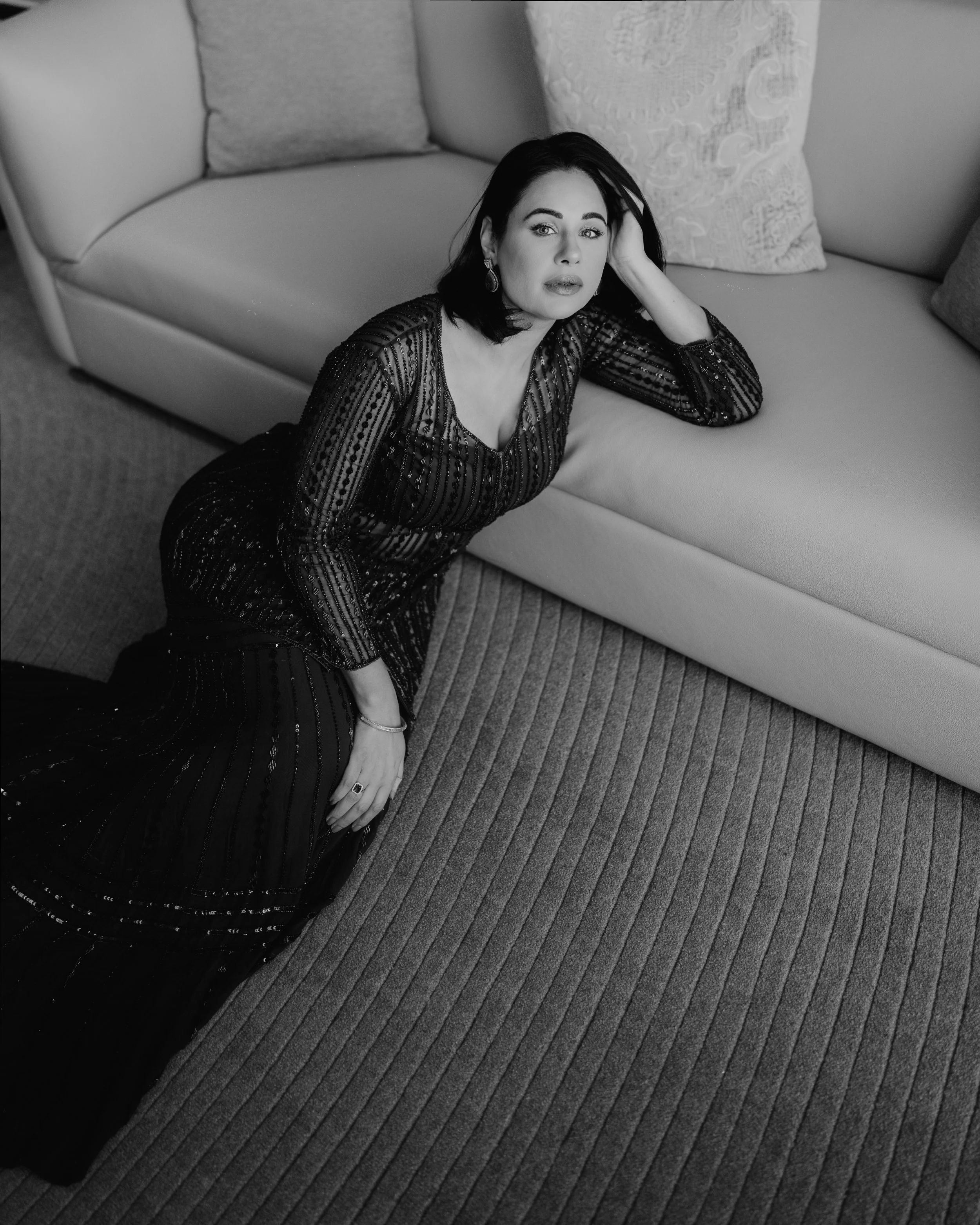 A woman with dark hair and earrings wearing a sheer, beaded dress; she is lying on her side on a patterned carpet next to a sofa with patterned cushions, looking into the camera.