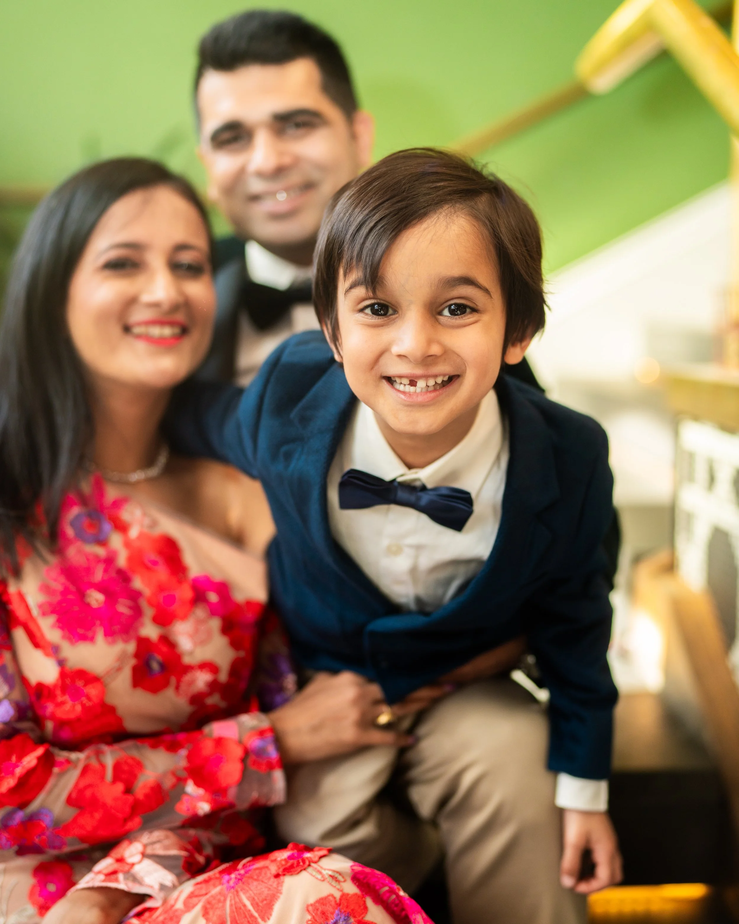 A young boy wearing a navy blazer and bow tie smiling as a woman in a colorful floral dress and a man in a tuxedo with a bow tie pose behind him.