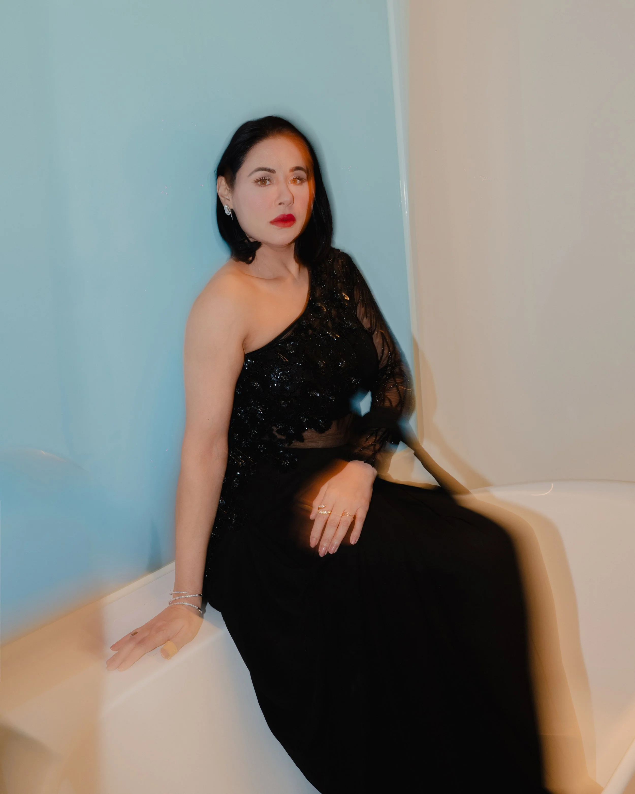 A woman with dark hair, red lipstick, and earrings sitting on a white surface against a blue and beige wall, wearing a black dress with lace and sequins.