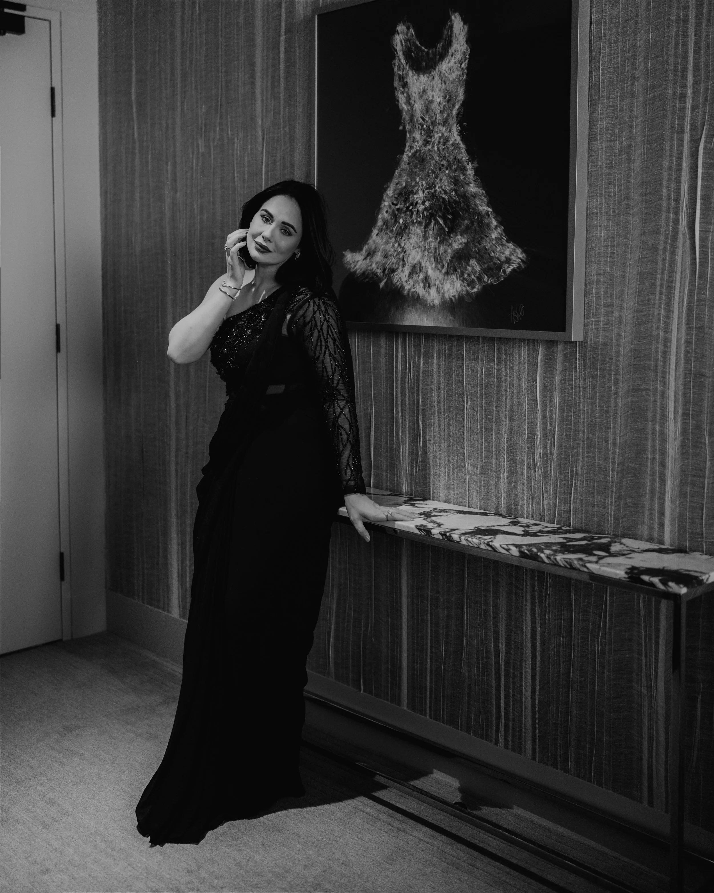 A woman with dark hair, wearing a long black dress with lace sleeves, stands by a marble-topped console in a modern interior, smiling slightly and touching her hair. Behind her, there is an abstract painting of a fox or wolf on a dark background, and