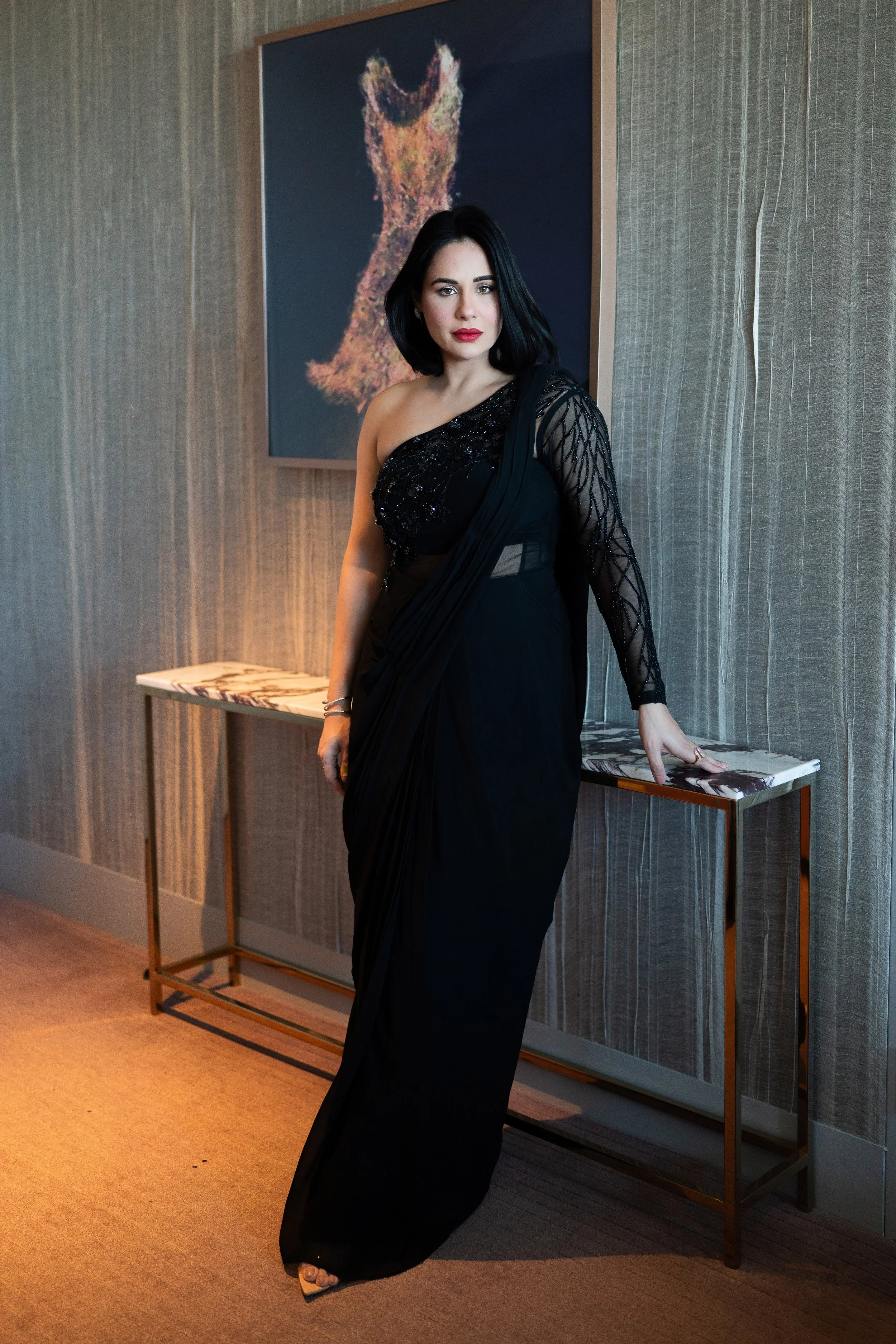 A woman in a black evening gown with one shoulder strap and lace detailing, standing in an art gallery near a marble-topped console table. Behind her is a large framed artwork of a dog made of colorful fragments. The woman has black hair and red lips