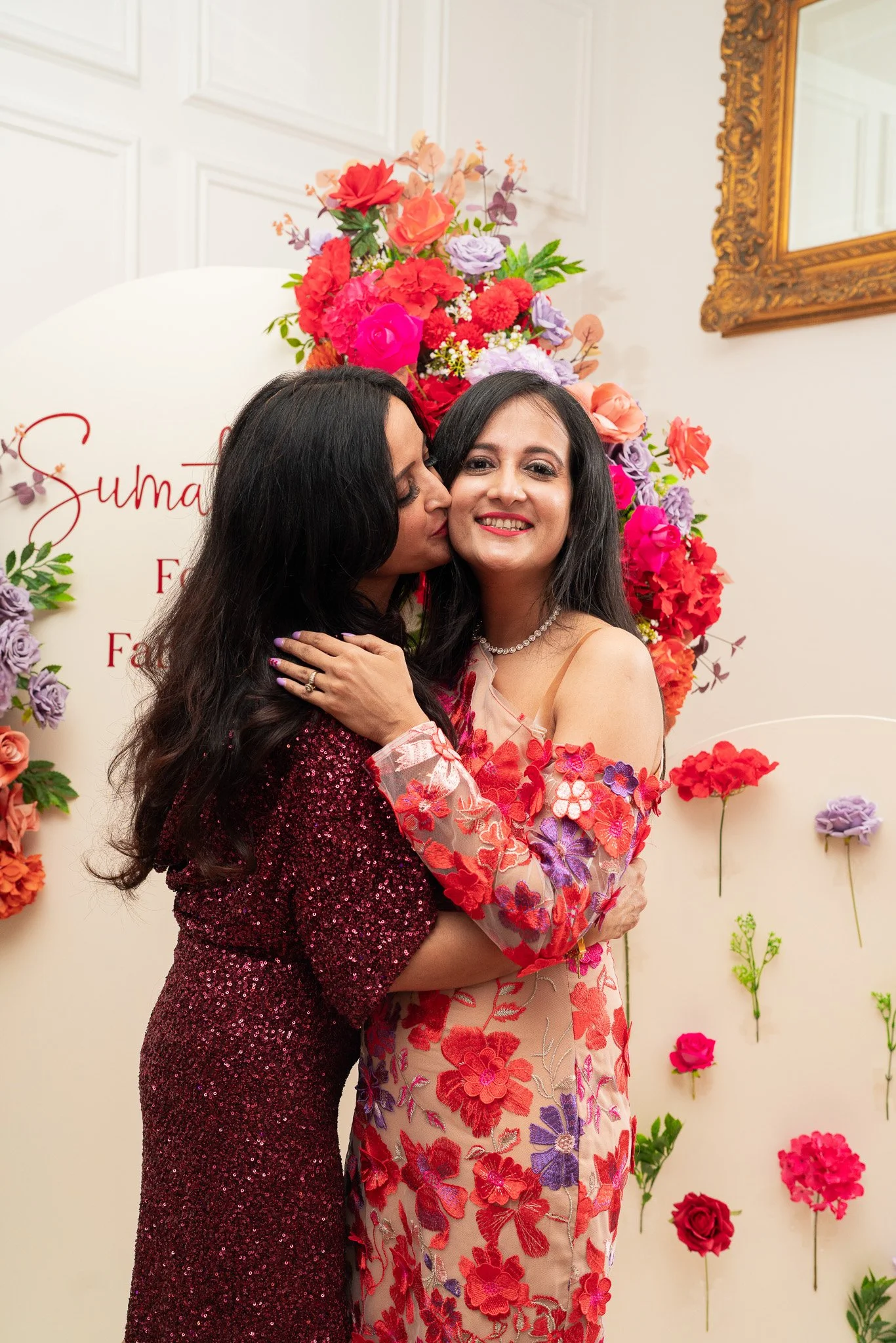 Two women celebrating at a floral-themed event. The woman on the right is smiling and wearing a beige dress with bold red, purple, and pink floral embroidery, and a pearl necklace. The woman on the left is wearing a dark red, sequined dress and is ki