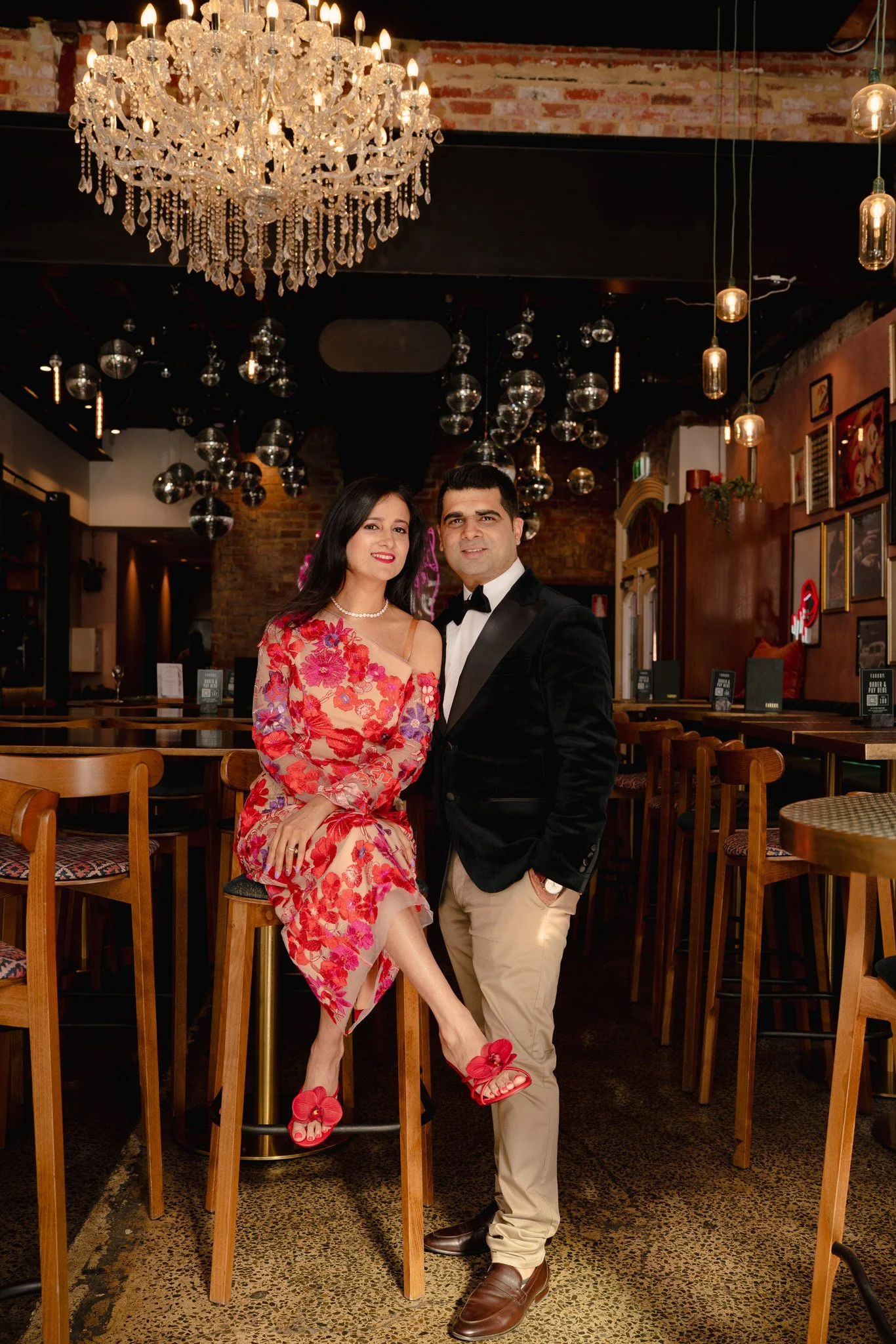 A couple dressed formally in an elegant restaurant. The woman is sitting on a high stool, wearing a floral pink dress with red high heels decorated with bows, and has dark hair. The man stands beside her with his hand in his pocket, wearing a black t