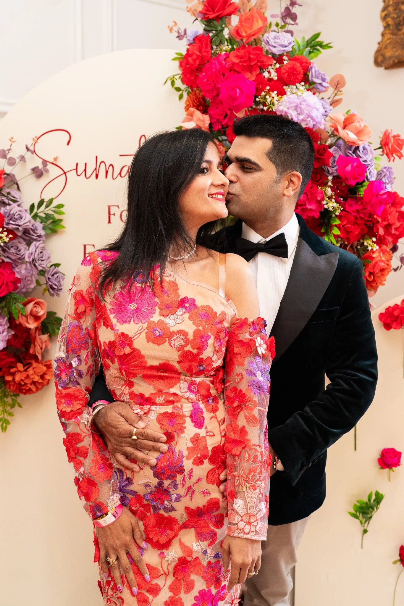 A smiling woman and a man in a tuxedo are standing close, with the woman holding her hand on her stomach. They are in front of a floral backdrop with a sign that says "Suma". The woman is wearing a colorful floral dress, and the man is in a black tux