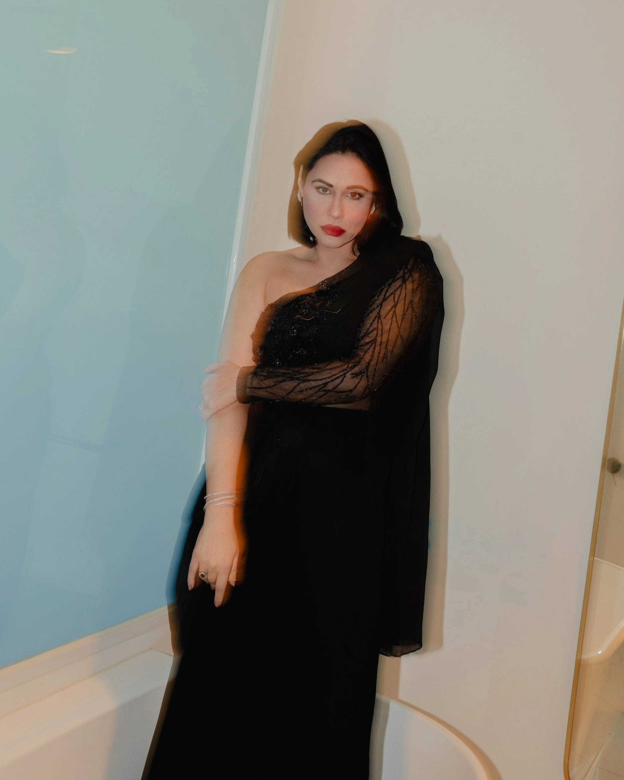 A woman posing against a wall, wearing a black dress with a sheer, embroidered sleeve, red lipstick, and earrings.