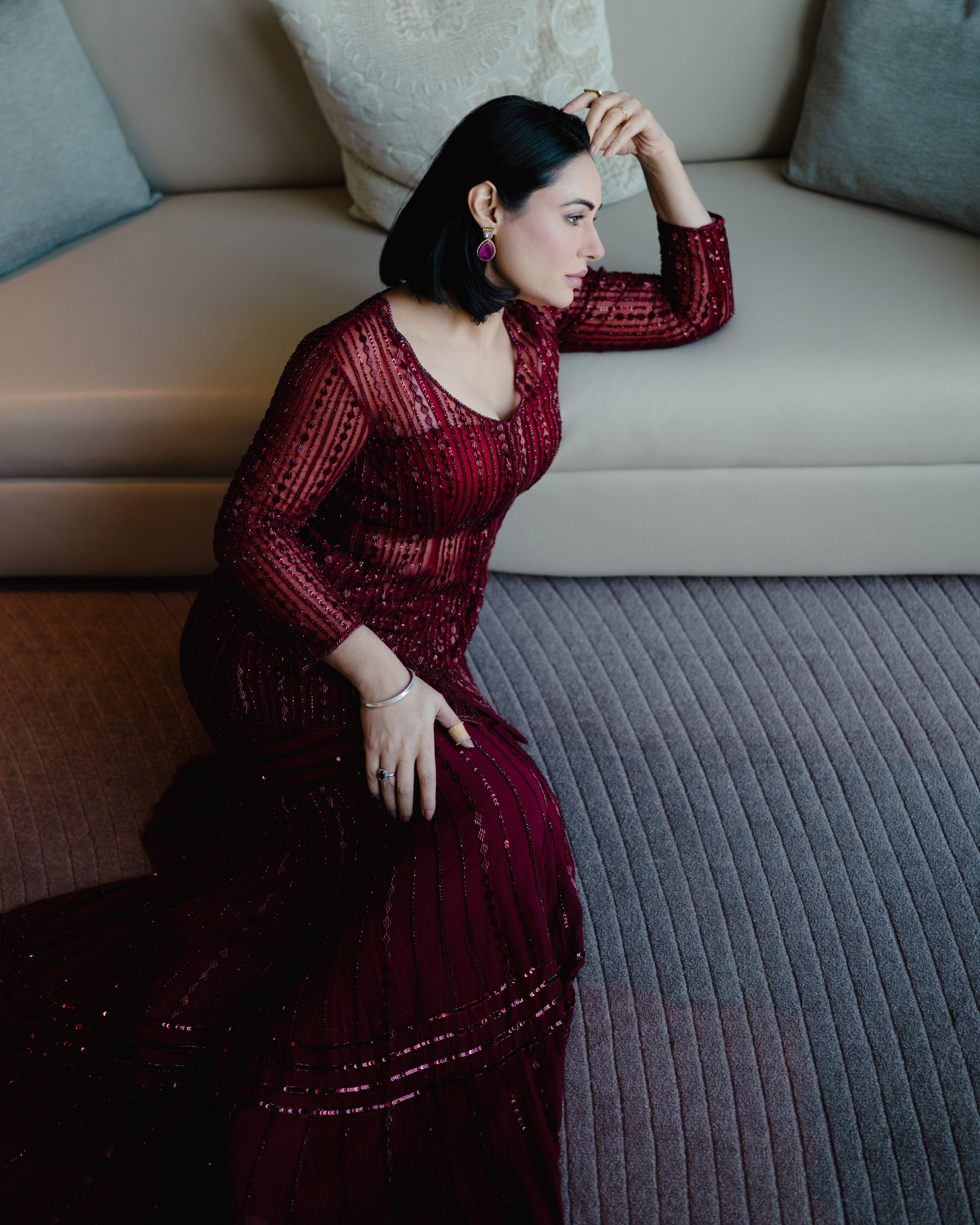 A woman with black hair and jewelry sitting on the floor with her head resting on a beige sofa with pillows, wearing a red beaded dress.