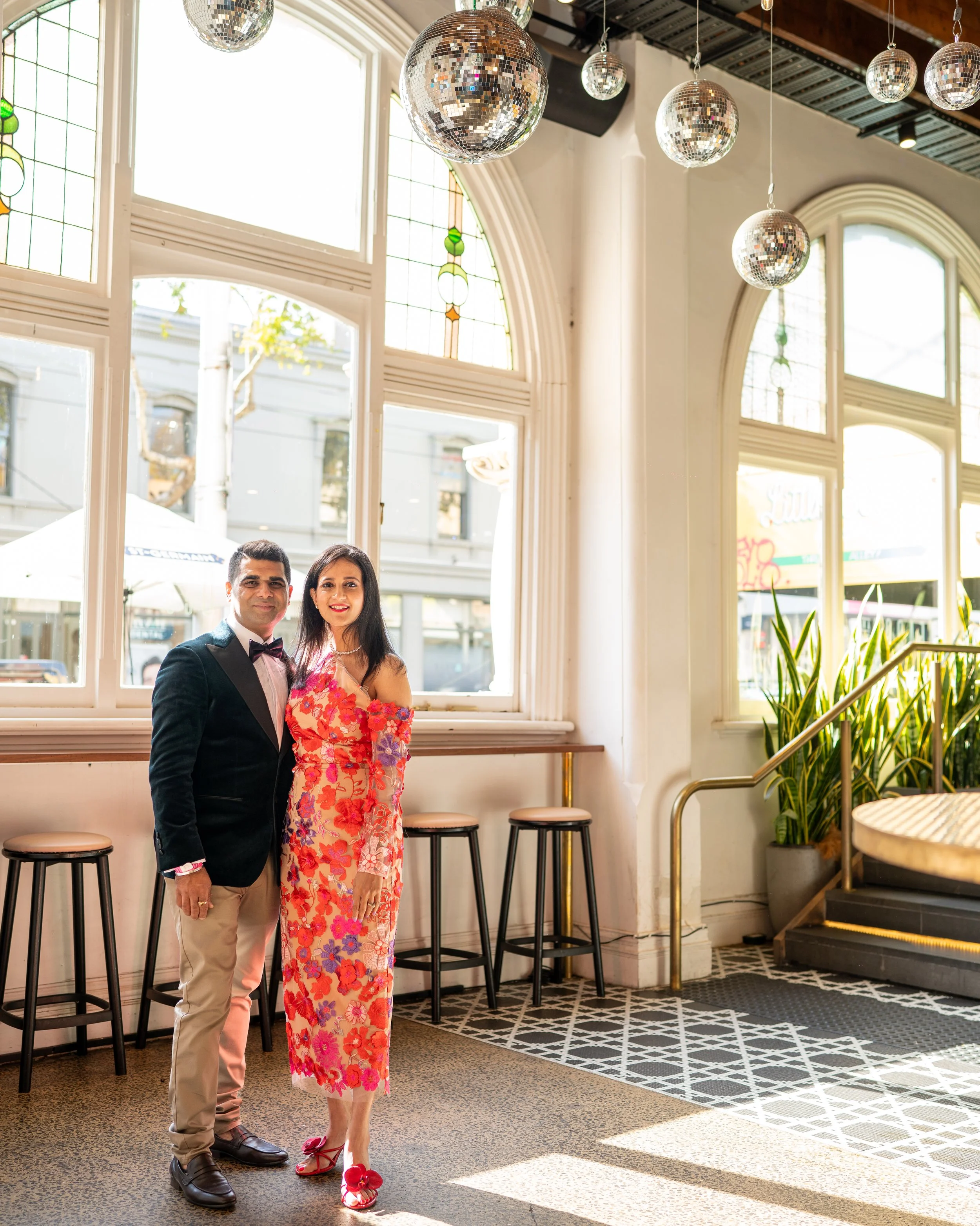 A man in a black tuxedo with a bow tie and beige trousers and a woman in a colorful floral dress standing inside a bright room with large windows, disco balls on the ceiling, bar stools along the window, and a staircase with railing.