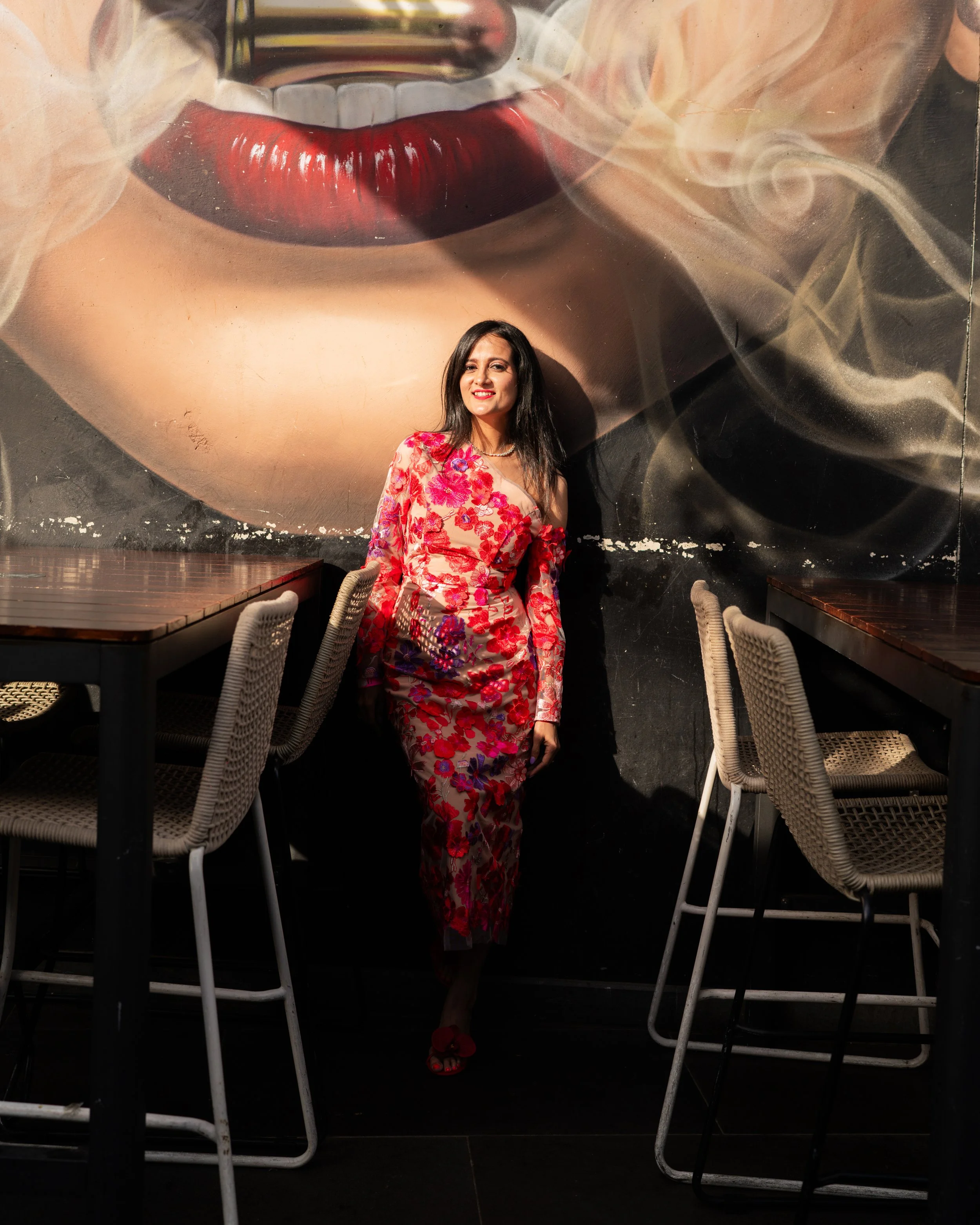 A woman with dark hair wearing a red floral dress and red shoes, standing in front of a graffiti mural of lips with white teeth and smoke around it, in a restaurant or cafe setting with chairs and tables.