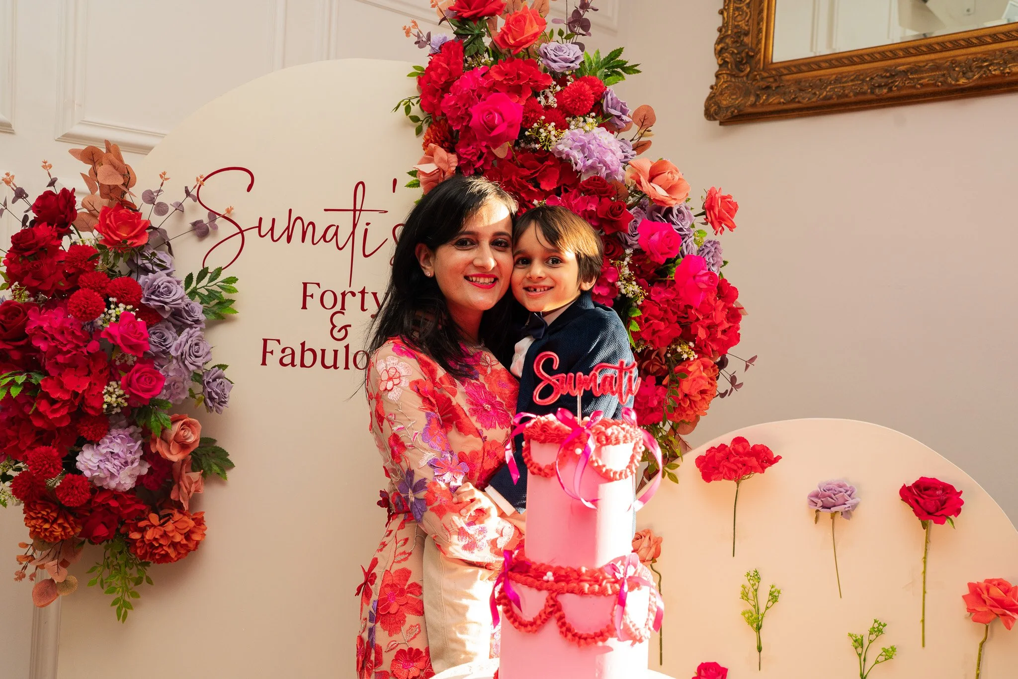 A woman and a young boy celebrating at a party with a pink cake and a floral backdrop.