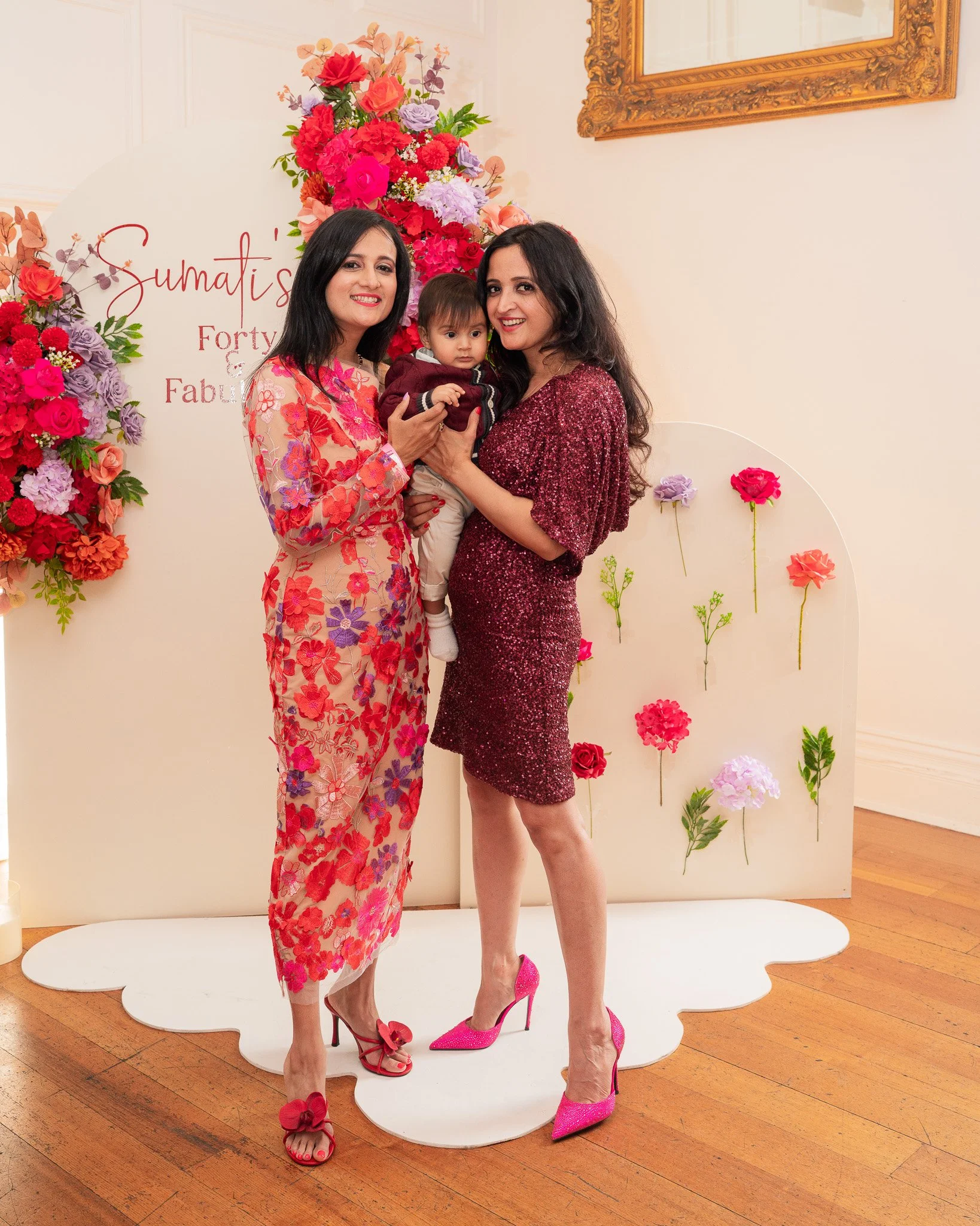 Two women and a young boy posing together at a celebration with a flower-themed backdrop. One woman wears a floral pink and red dress, a woman wears a sparkly red dress, and the boy is dressed in a maroon jacket. They are all smiling, and the setting