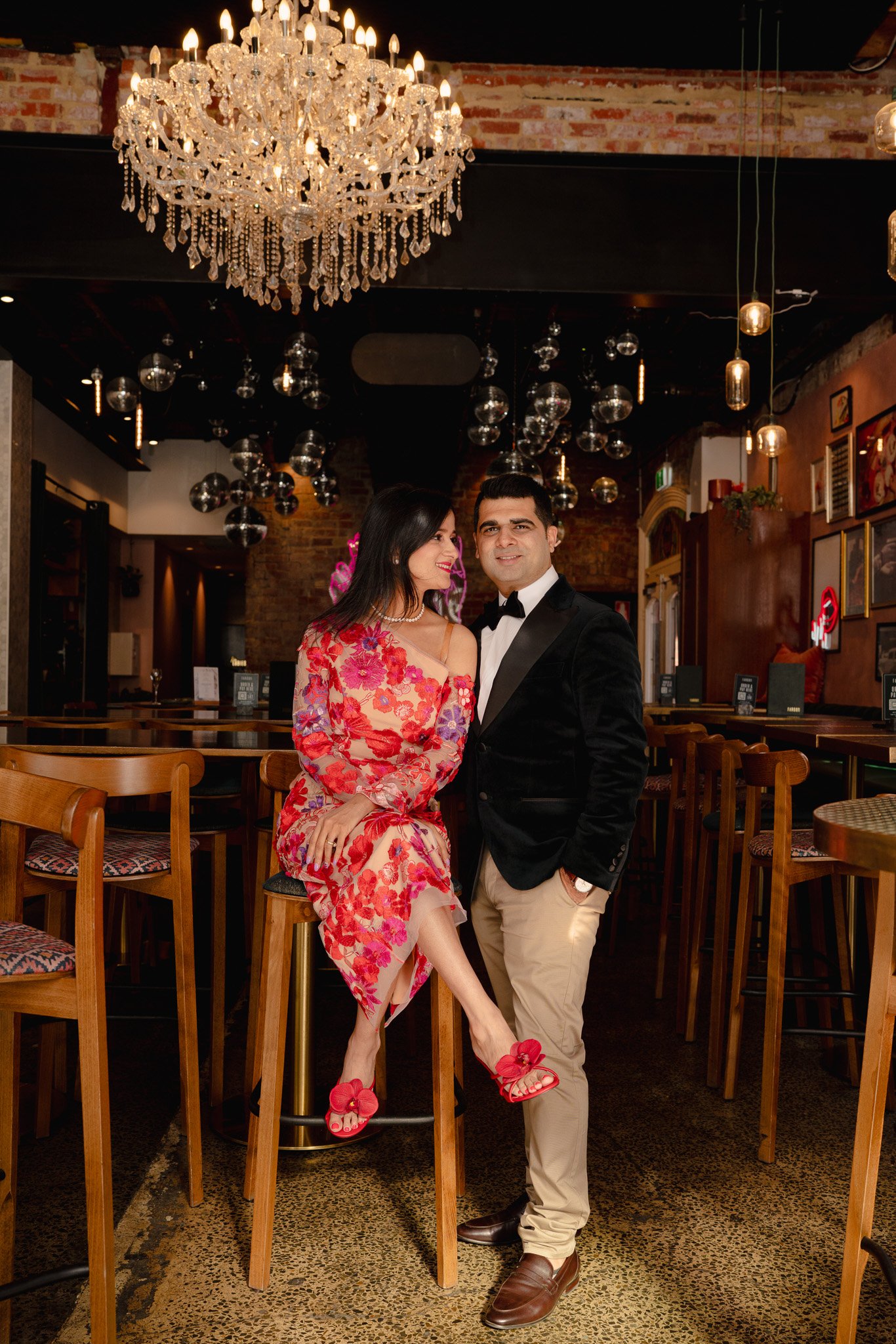 A man in a black tuxedo and a woman in a colorful floral dress posing together inside a dimly lit restaurant with a large chandelier above them and numerous hanging light bulbs.