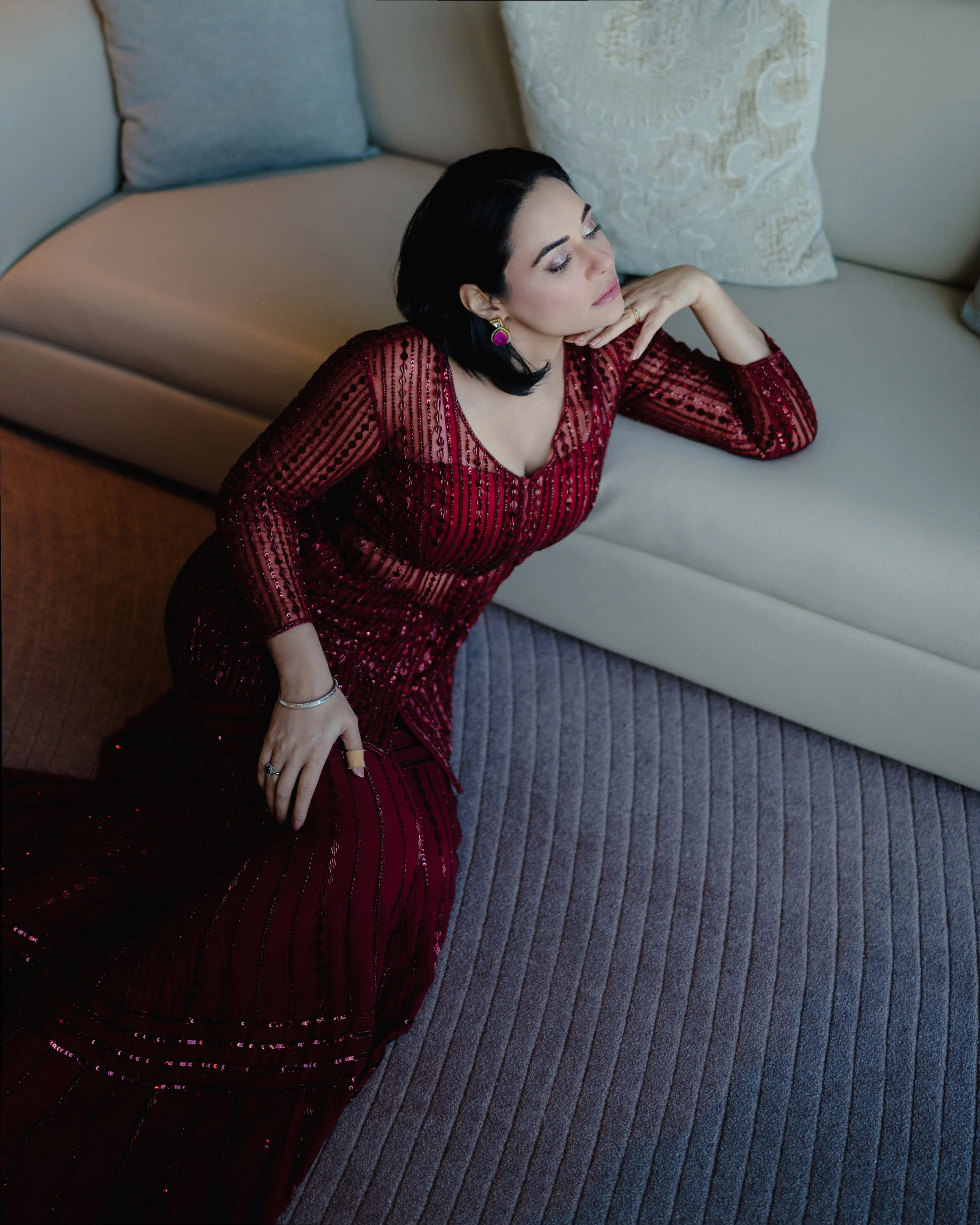 A woman with dark hair, makeup, and earrings is wearing a red dress with black stripes and sequins, posing with her hand near her chin while sitting on a beige couch with decorative pillows and a striped carpet underneath.