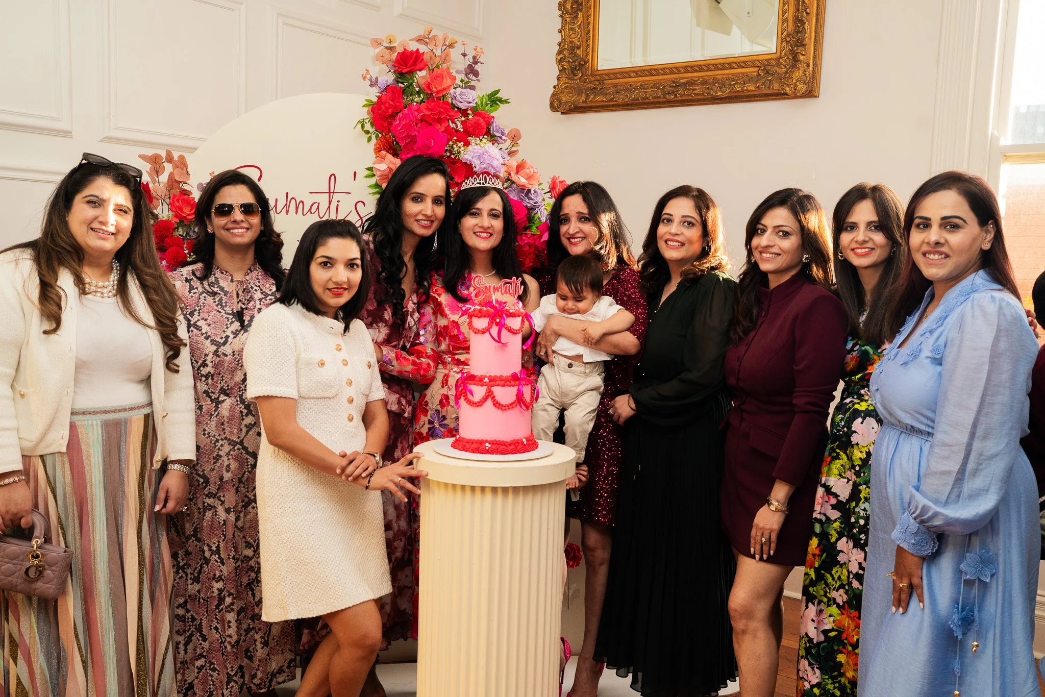 A group of women and a young boy celebrating a birthday, standing around a tall pink cake with a crown on top, in front of a large floral arrangement and a wall with a mirror, with some people smiling and others looking at the camera.