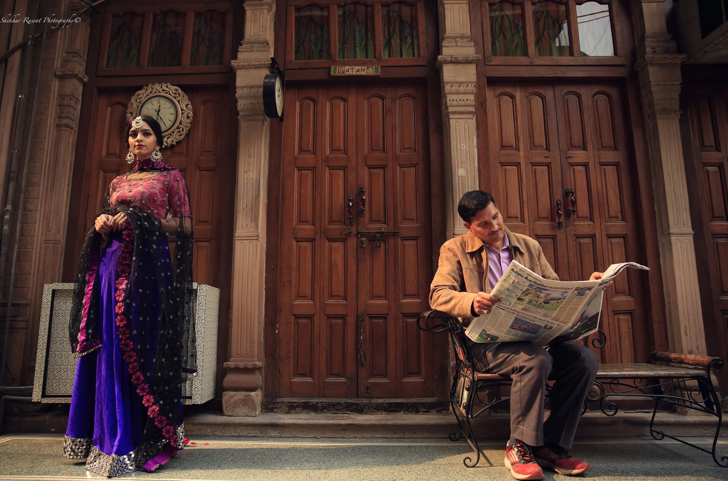 Woman in traditional Indian attire beside a man reading a newspaper at a heritage doorway, storytelling portrait by Melbourne photographer Shekhar Rawat