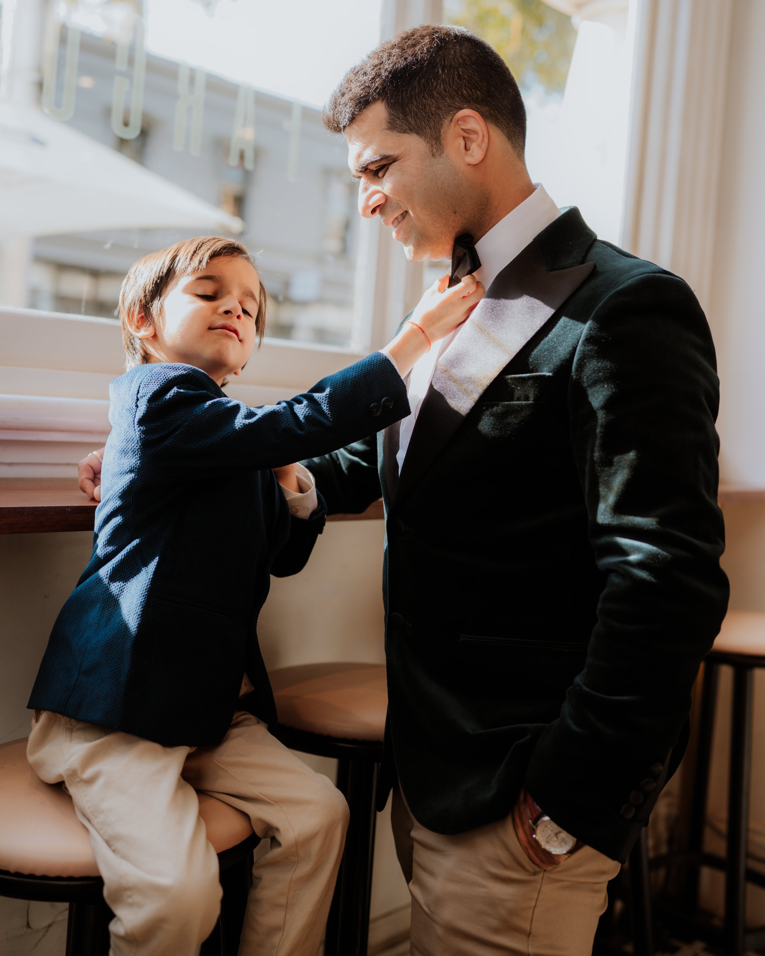 A man in a tuxedo adjusting his bow tie while a boy in a blazer sits and looks at him.