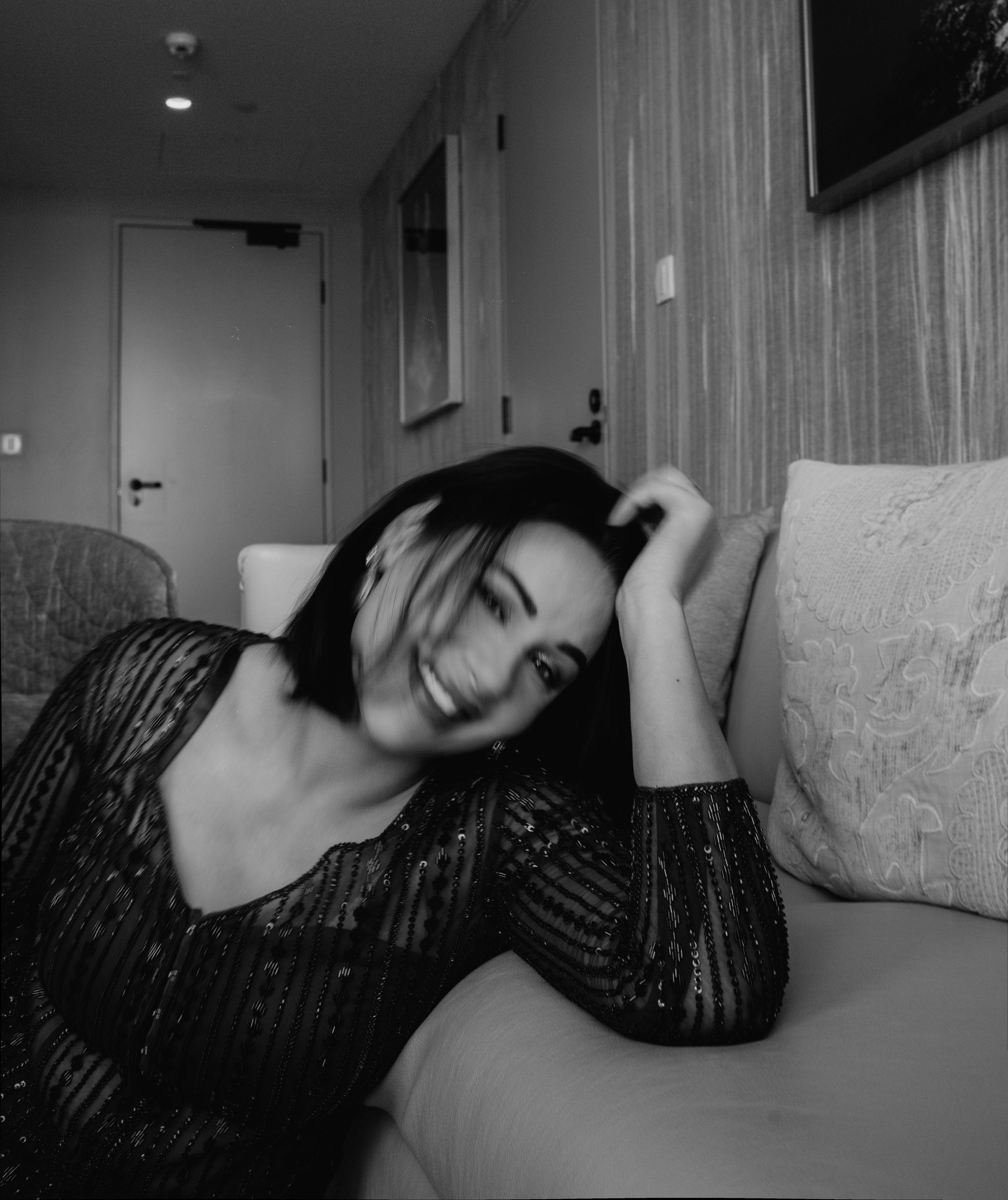 A woman with dark hair and a striped dress sitting on a bed, smiling and posing for the camera in a bedroom.