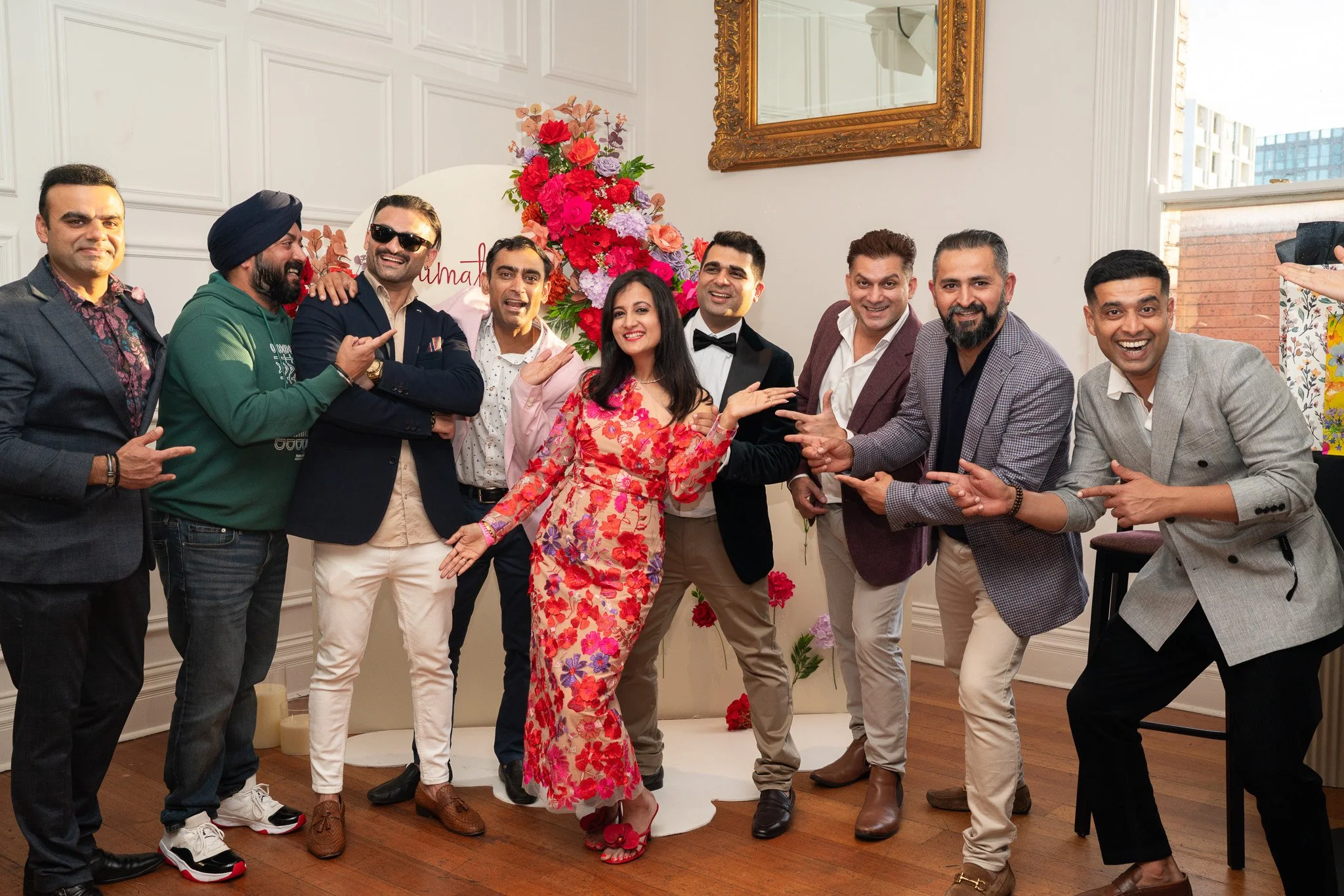 A group of ten people, including women and men of diverse ethnicities, are smiling and posing together in a room with white walls and a large framed mirror. They are dressed in formal and semi-formal attire, with some pointing at each other or making