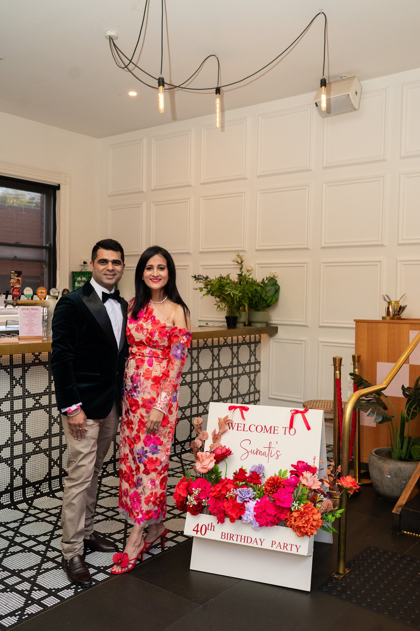 A man in a black tuxedo and a woman in a pink floral dress standing at a 40th birthday party celebration. There is a white sign with red ribbons that reads, "Welcome to Sumati's 40th Birthday Party," surrounded by colorful flowers. The background fea