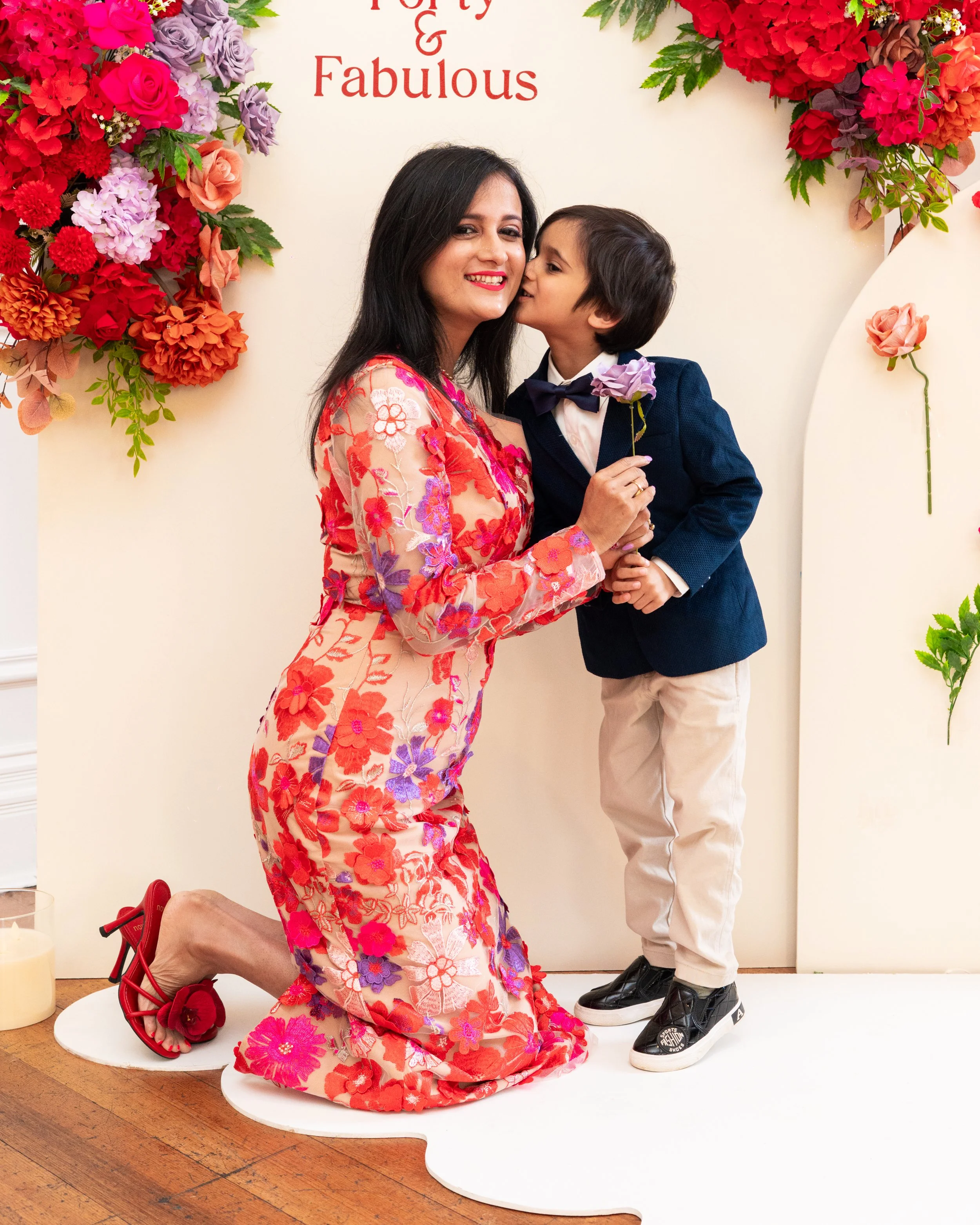 A woman kneels on a white platform, wearing a colorful floral dress and red high heels, with a man standing behind her, kissing her cheek. The man wears a navy blazer, white shirt, beige pants, and black sneakers. There are colorful flowers decoratin