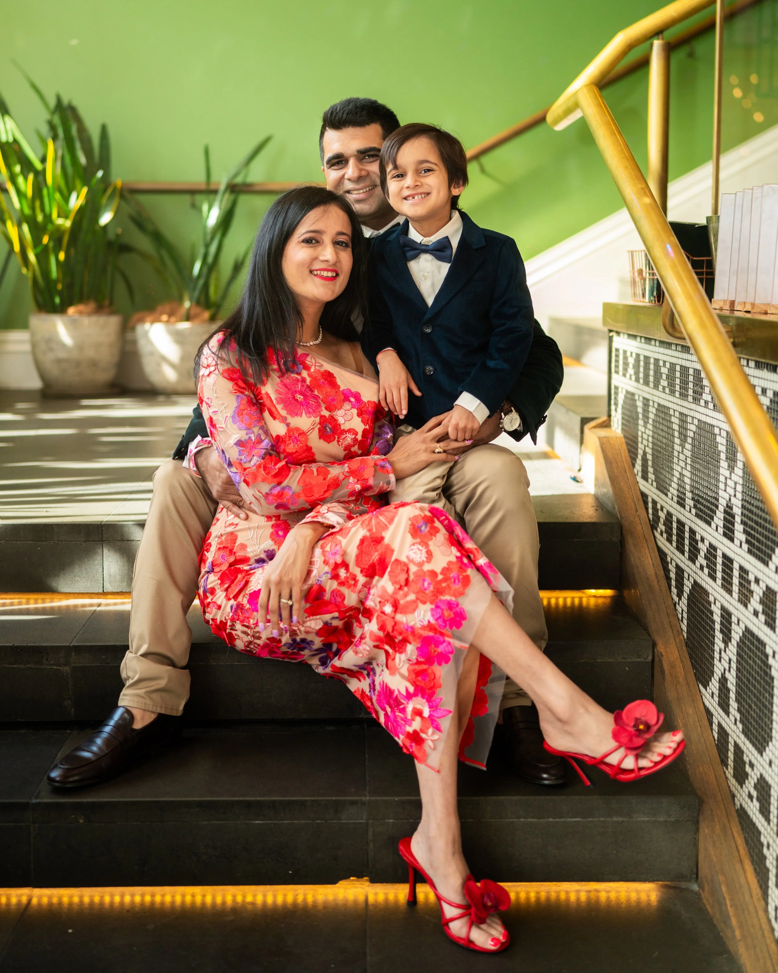 A family of three posing on indoor black stairs with a green wall background. The woman is wearing a pink and red floral dress, red heels, and has black hair. The man is wearing beige pants, black shoes, and a dark blazer. The boy is dressed in a dar