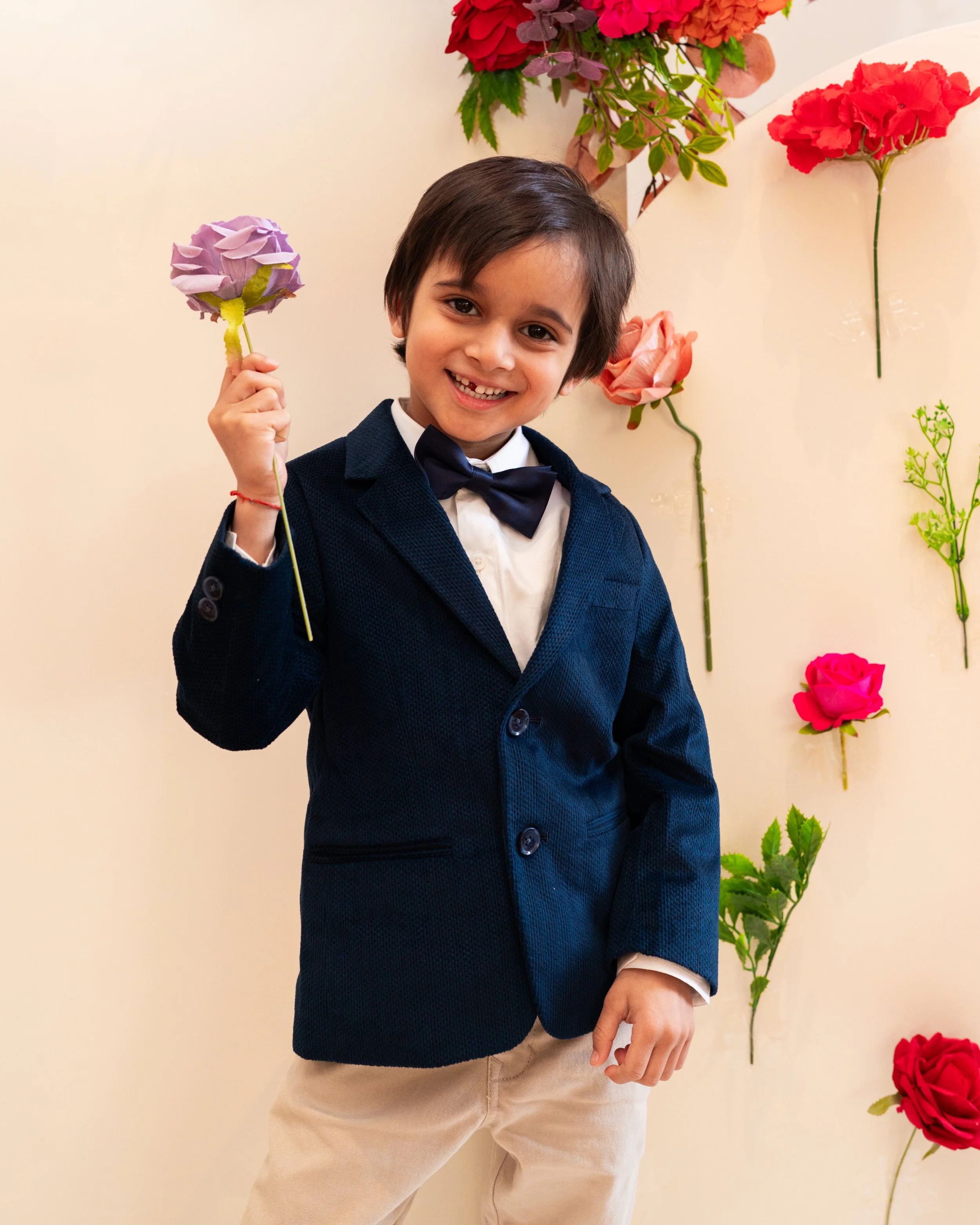 A young boy with dark hair is smiling and holding a purple flower while standing in front of a light-colored wall decorated with pink, red, and orange artificial flowers.