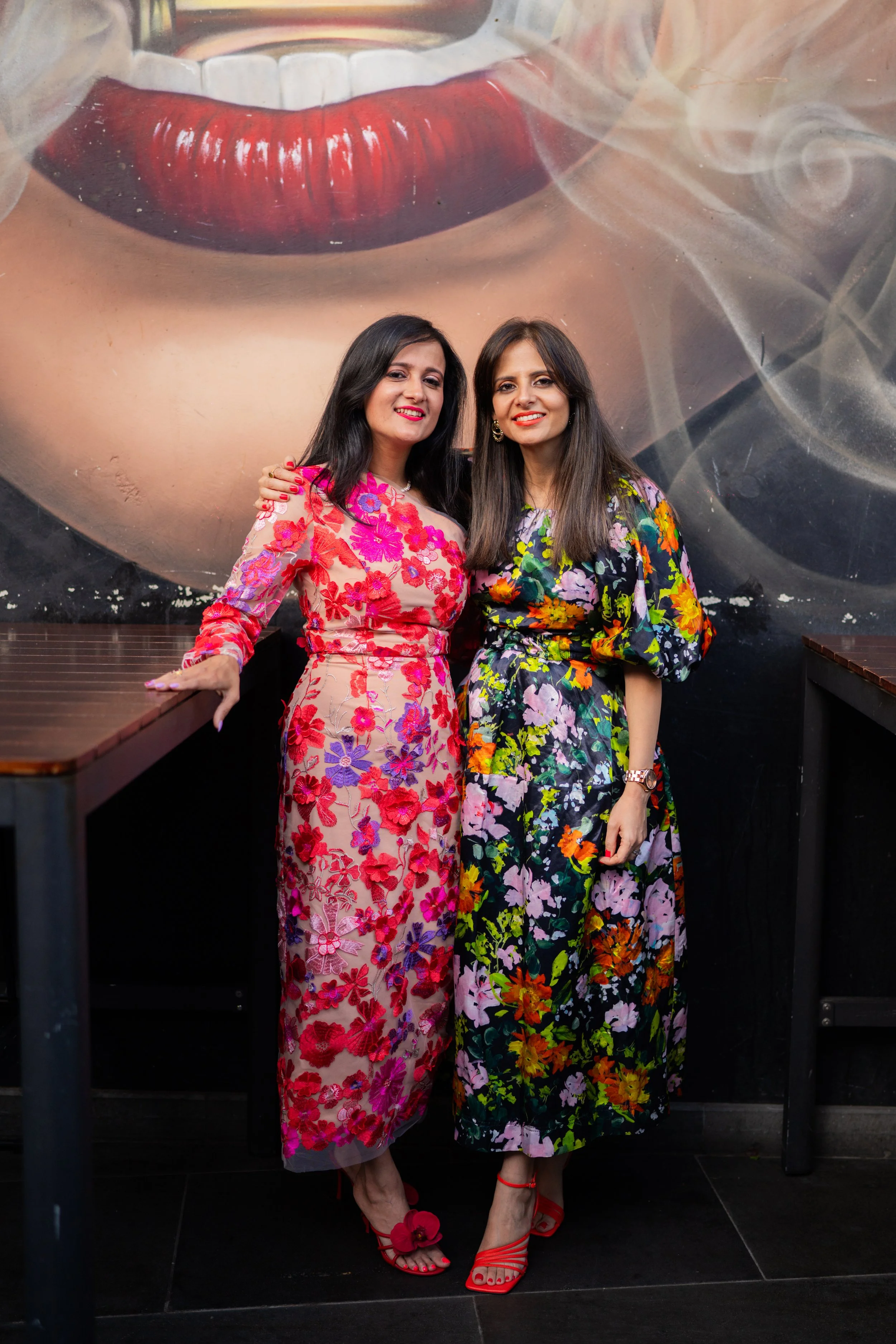 Two women are standing close together in front of a large mural of a woman's face with red lips, wearing colorful floral dresses and red heels. One woman has her hand on the other's shoulder and they are smiling.