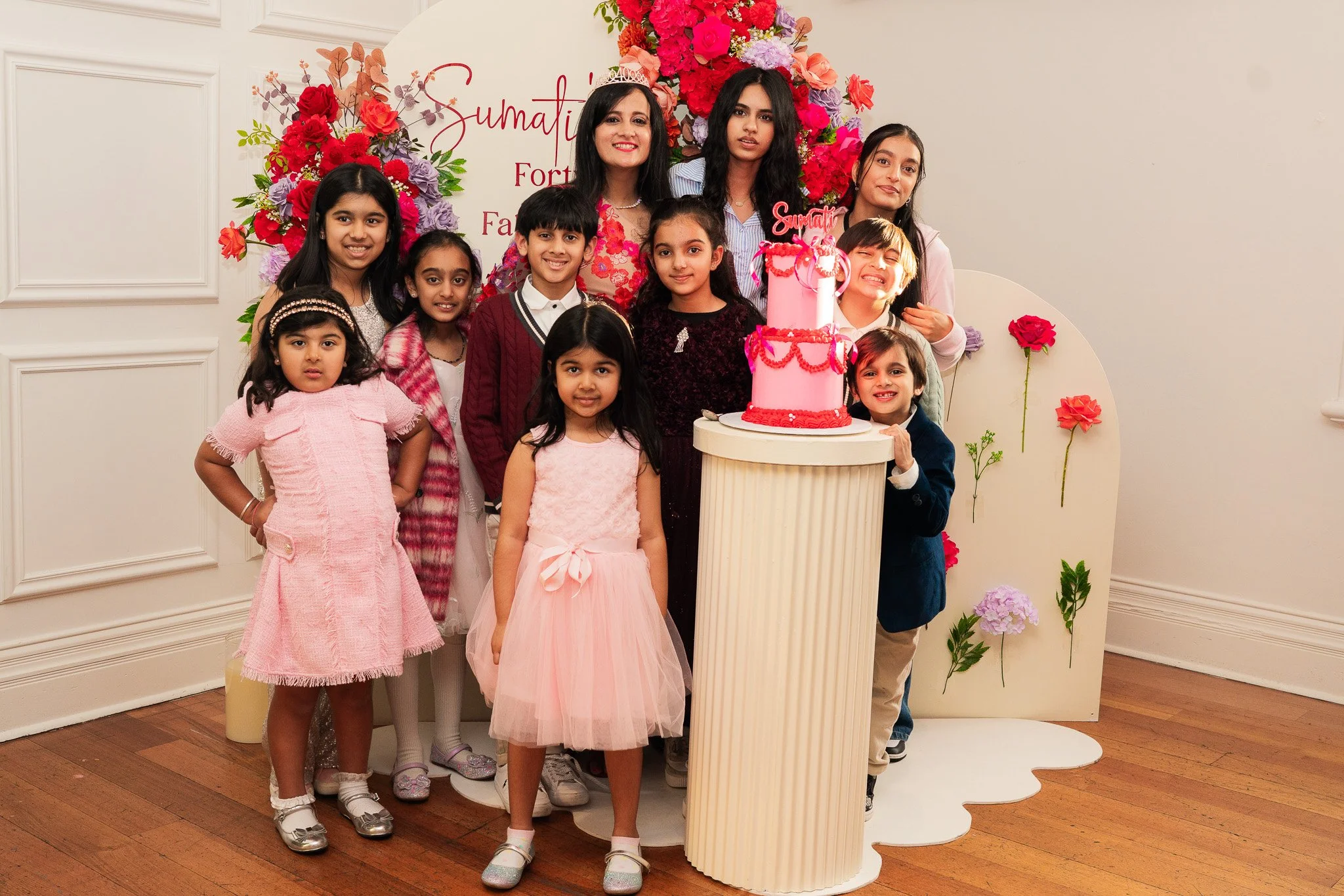 A group of ten children and one woman standing together at a birthday celebration with a large pink cake decorated with red and pink flowers and a sign that says "Sumati" in cursive. They are all smiling and dressed in colorful clothes, with some gir