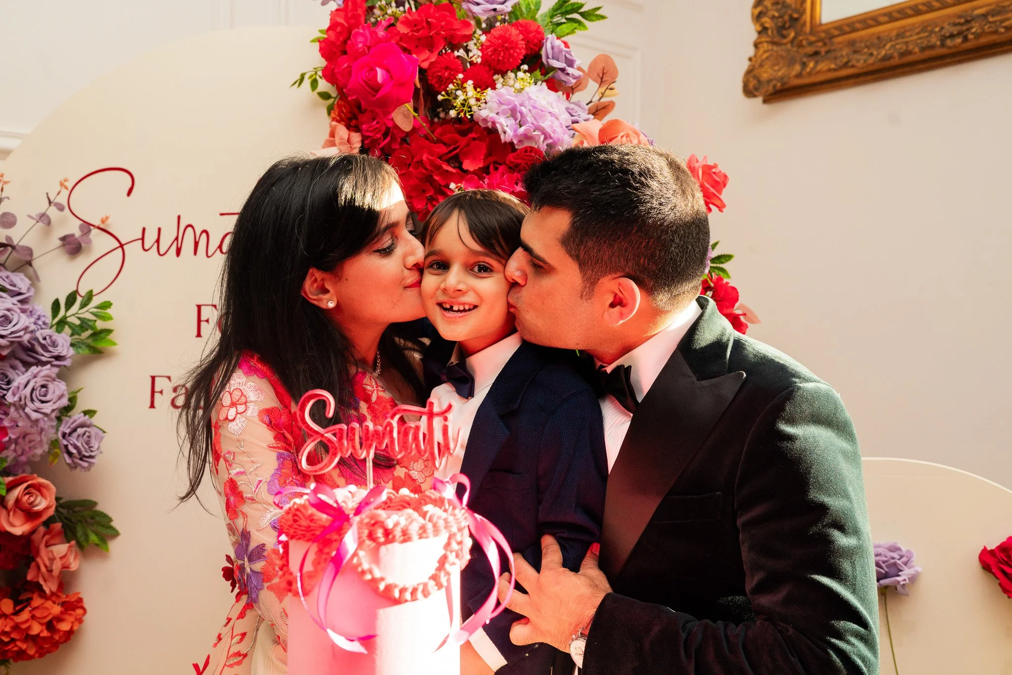 A family of three celebrating a birthday with a pink cake decorated with flowers. The mother and father are kissing their smiling young son on the cheeks. Behind them is a floral backdrop and a sign that reads "Sundari".
