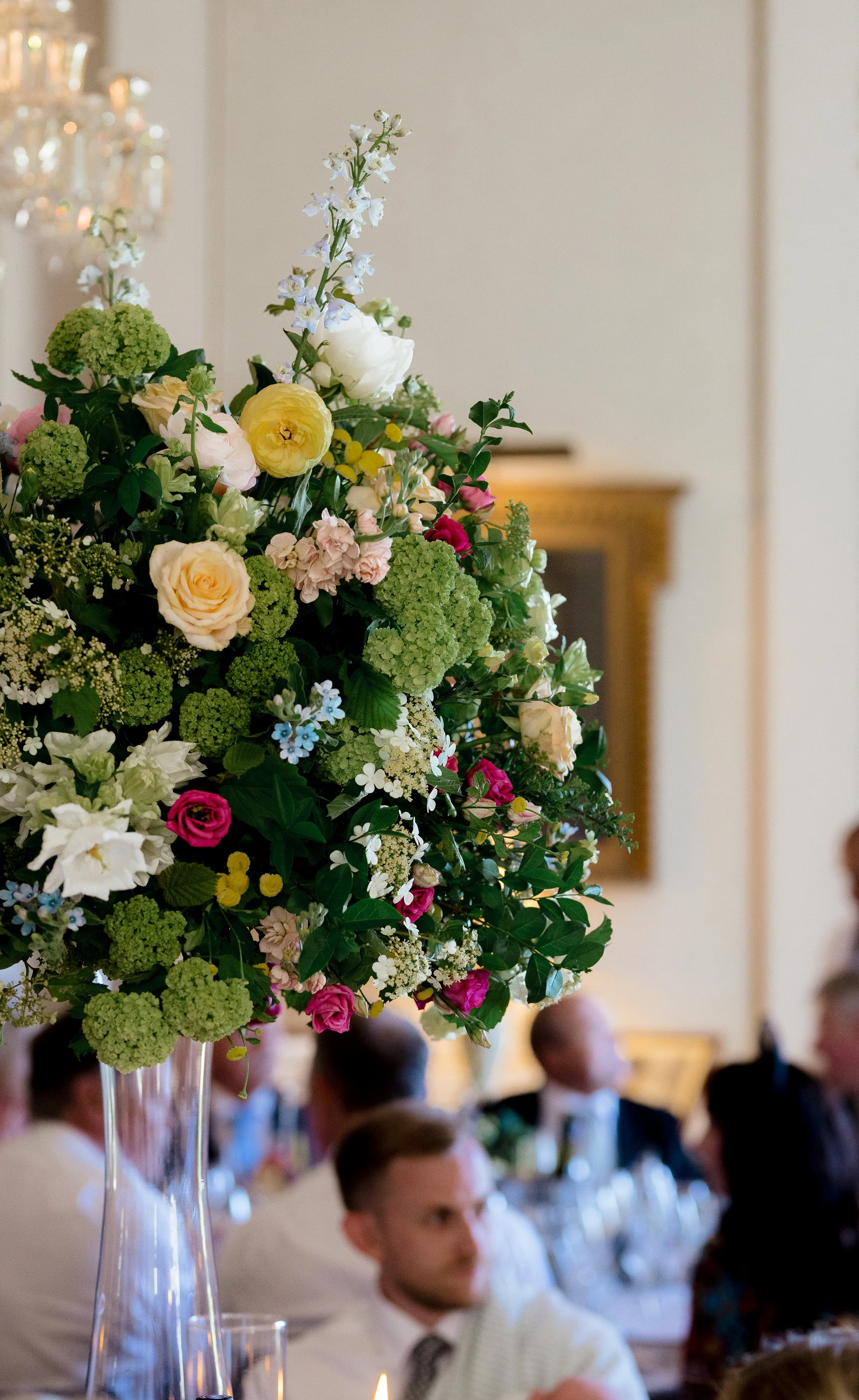 A large floral arrangement with white, pink, yellow, and green flowers on a tall glass vase at a formal event, with blurred guests in the background.