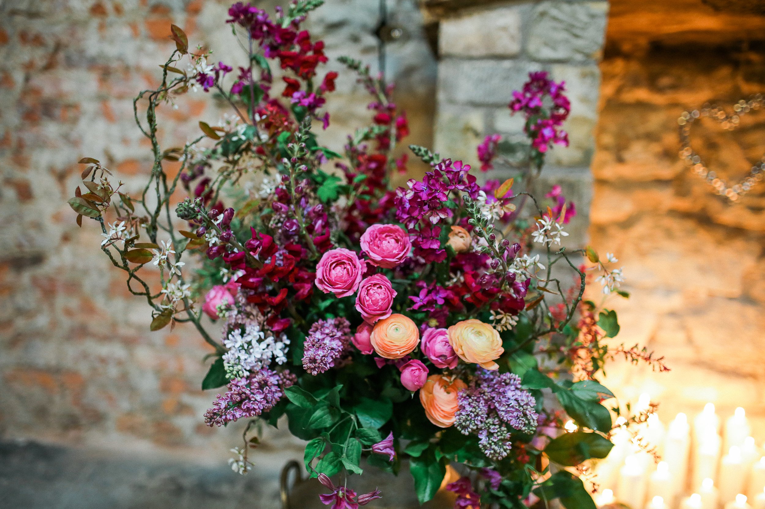 Vibrant floral arrangement with various pink, purple, and orange flowers, set against a rustic brick wall background, with soft candlelight illumination.