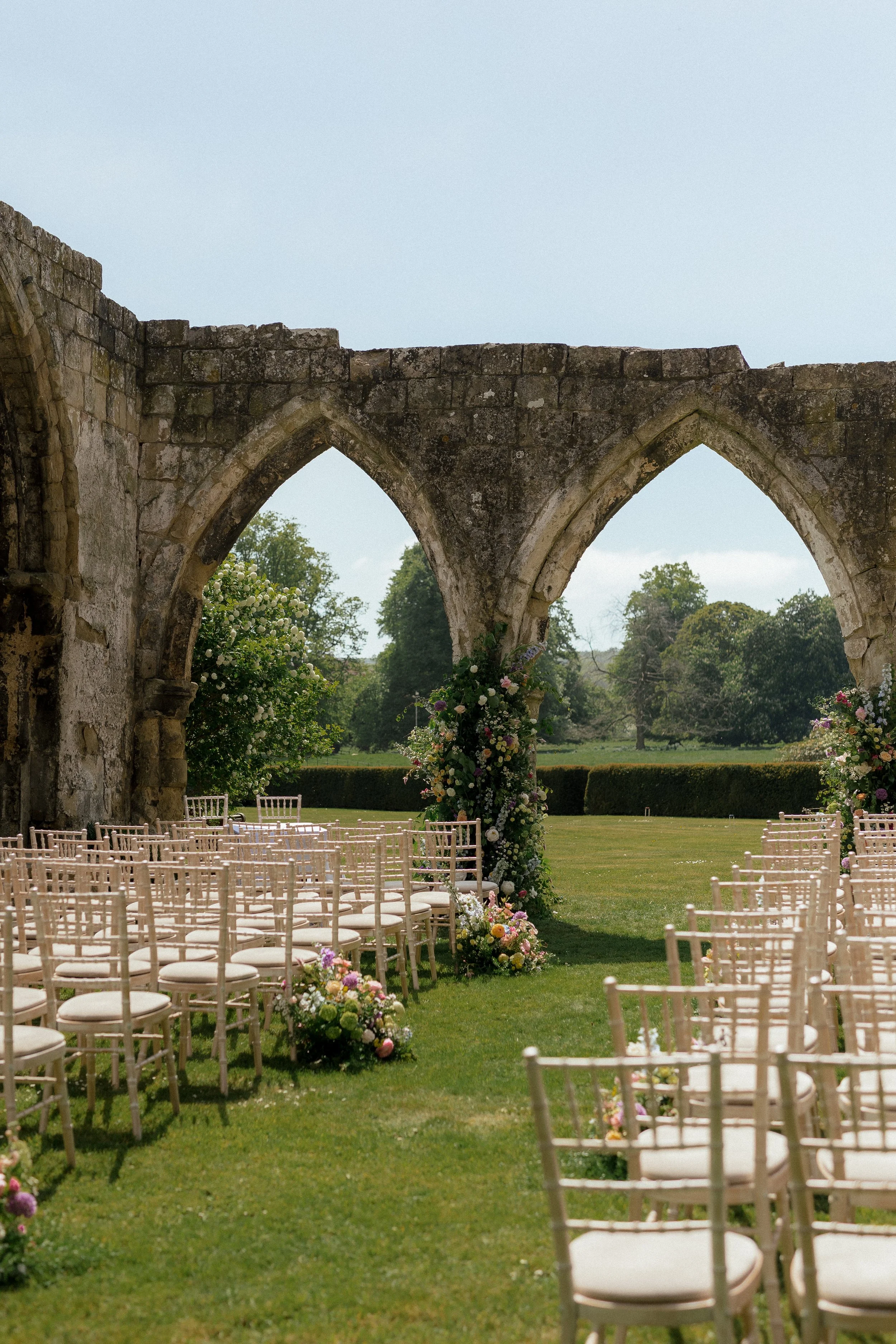 Outdoor wedding setup with white chairs and floral arrangements under stone arches on a grassy field.